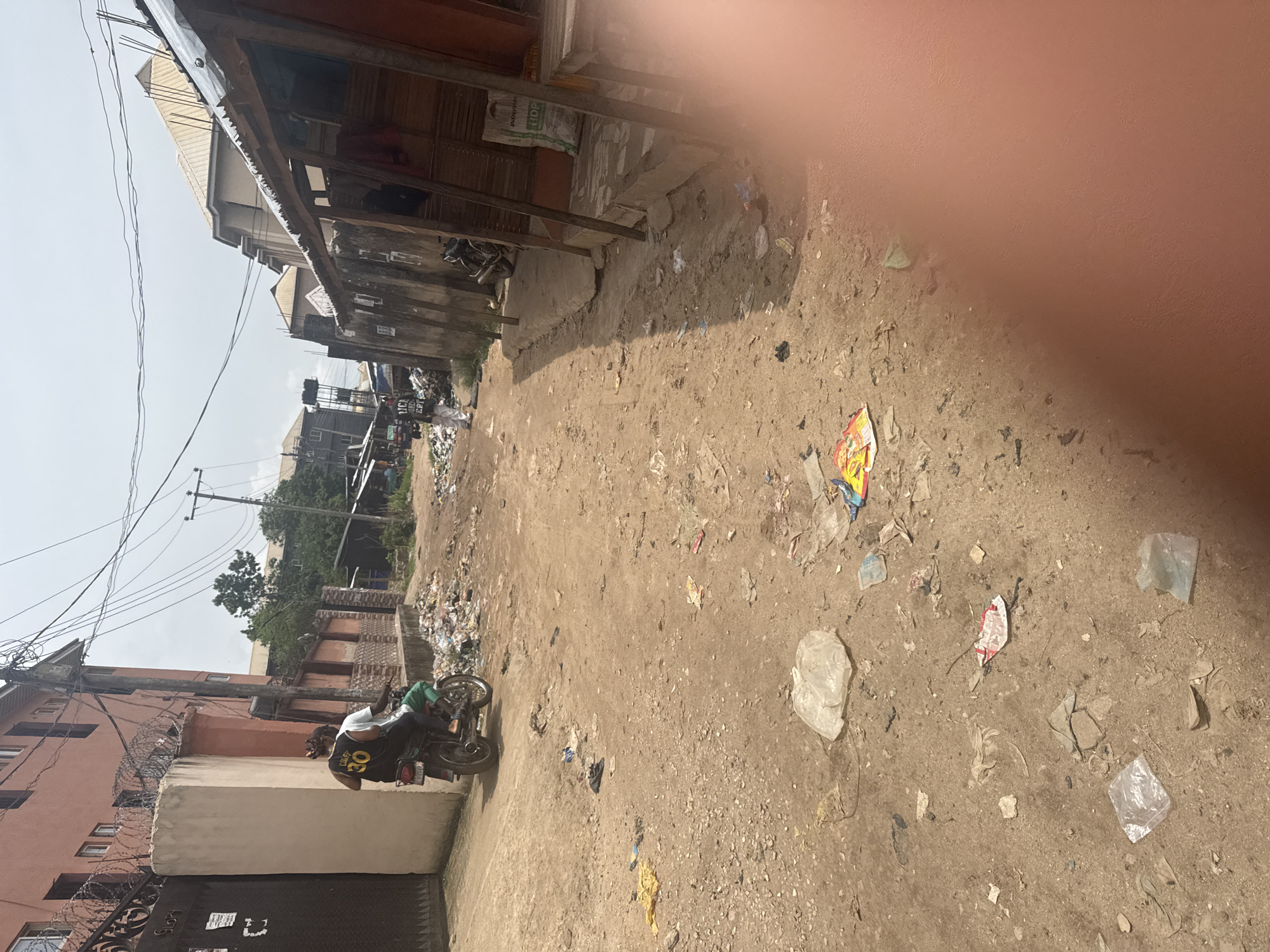 This vertical image captures a dusty street scene in Ihiagwa, Nigeria, under bright daylight. The unpaved road, made of packed dirt and gravel, is extensively littered with various pieces of debris, including plastic bags, wrappers, and discarded packaging materials.
In the midground, a man wearing a black t-shirt with "COL.07 30" visible on the back is seated on a green and black motorcycle, facing away from the viewer. To the left, behind the man, stands a multi-story building with an orange-brown facade, partially obscured by a grey wall and a black metal gate topped with barbed wire. Utility poles and a network of power lines are visible against the bright, clear sky.
Along the right side of the street, a row of single and multi-story structures with corrugated metal roofs lines the road. One building features a dark, weathered exterior, and a white sack with "INDORAM HDP" printed on it rests near its base. Further down the street, another individual can be vaguely discerned amidst a larger pile of accumulated waste.
The image conveys a clear, sunny day. A large, blurred reddish-pink area, likely a finger or hand, obstructs the upper right portion of the original photograph.