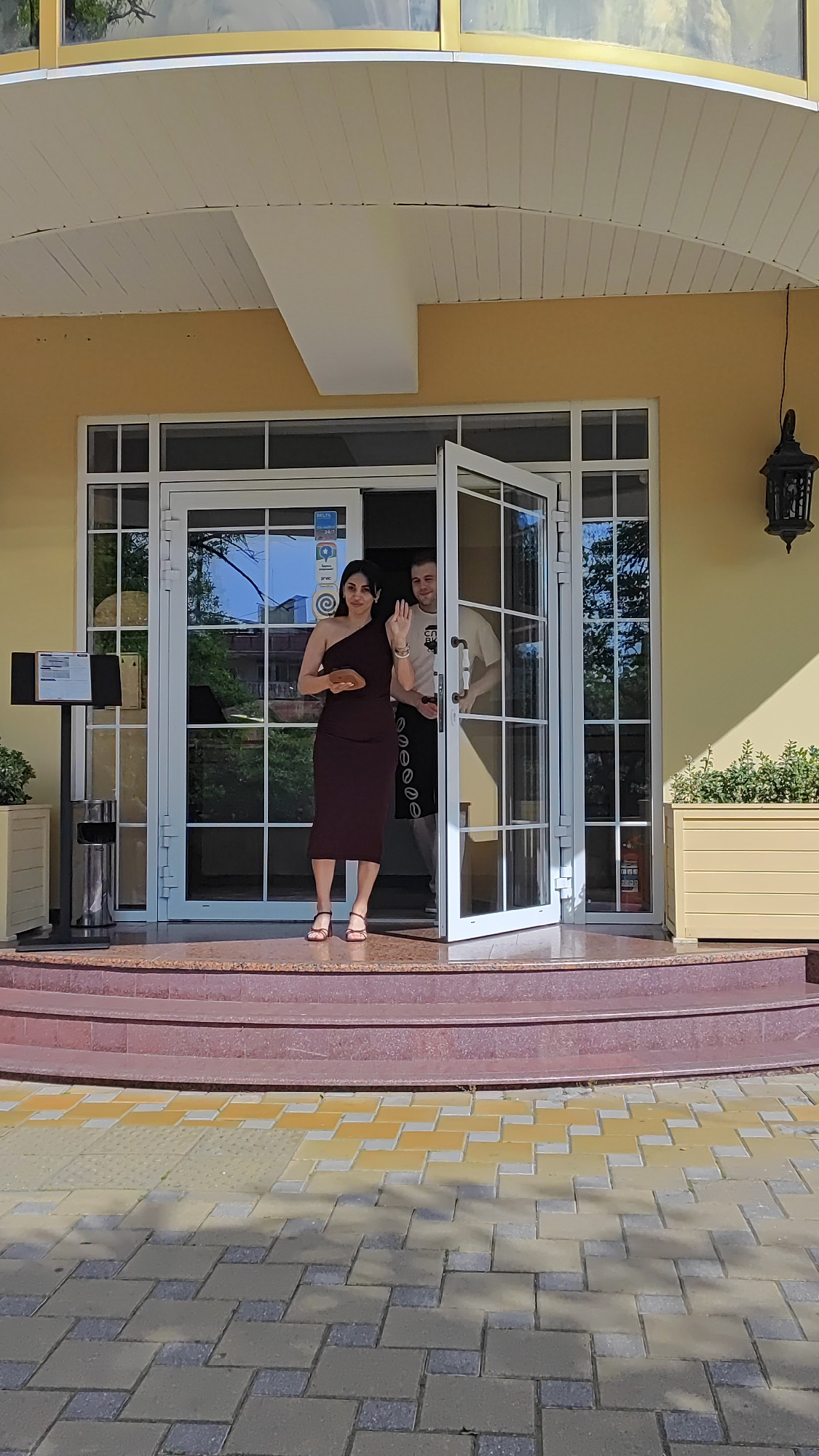 A woman in a maroon dress stands on the front steps of a building and waves. She holds a small, light brown object in her left hand. Behind her, a man in a light-colored t-shirt and dark apron stands in a doorway. The building appears to be a restaurant or cafe with large glass doors and a yellow exterior. Planters with greenery are visible on either side of the entrance. The steps are made of reddish-brown material, and the walkway in front is paved with interlocking stones in yellow and gray. The scene is outdoors, under a covered entryway.