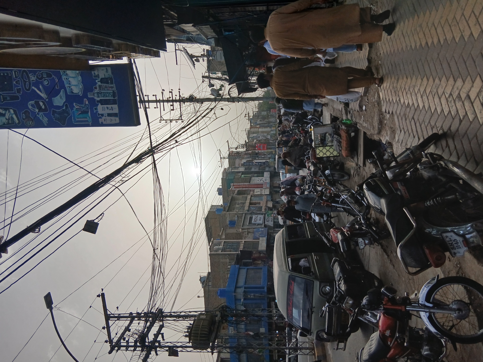 The image depicts a bustling street scene in Quetta City Tehsil, Pakistan. The street is lined with buildings, many of which have signage in Urdu or English. Overhead, a dense network of electrical wires crisscrosses the sky. The street itself is crowded with motorcycles parked along the sides, and a light-colored van is visible in the middle of the road. People are walking along the sidewalk, some wearing traditional Pakistani clothing, such as shalwar kameez. The overall atmosphere suggests a busy commercial area during daylight hours.