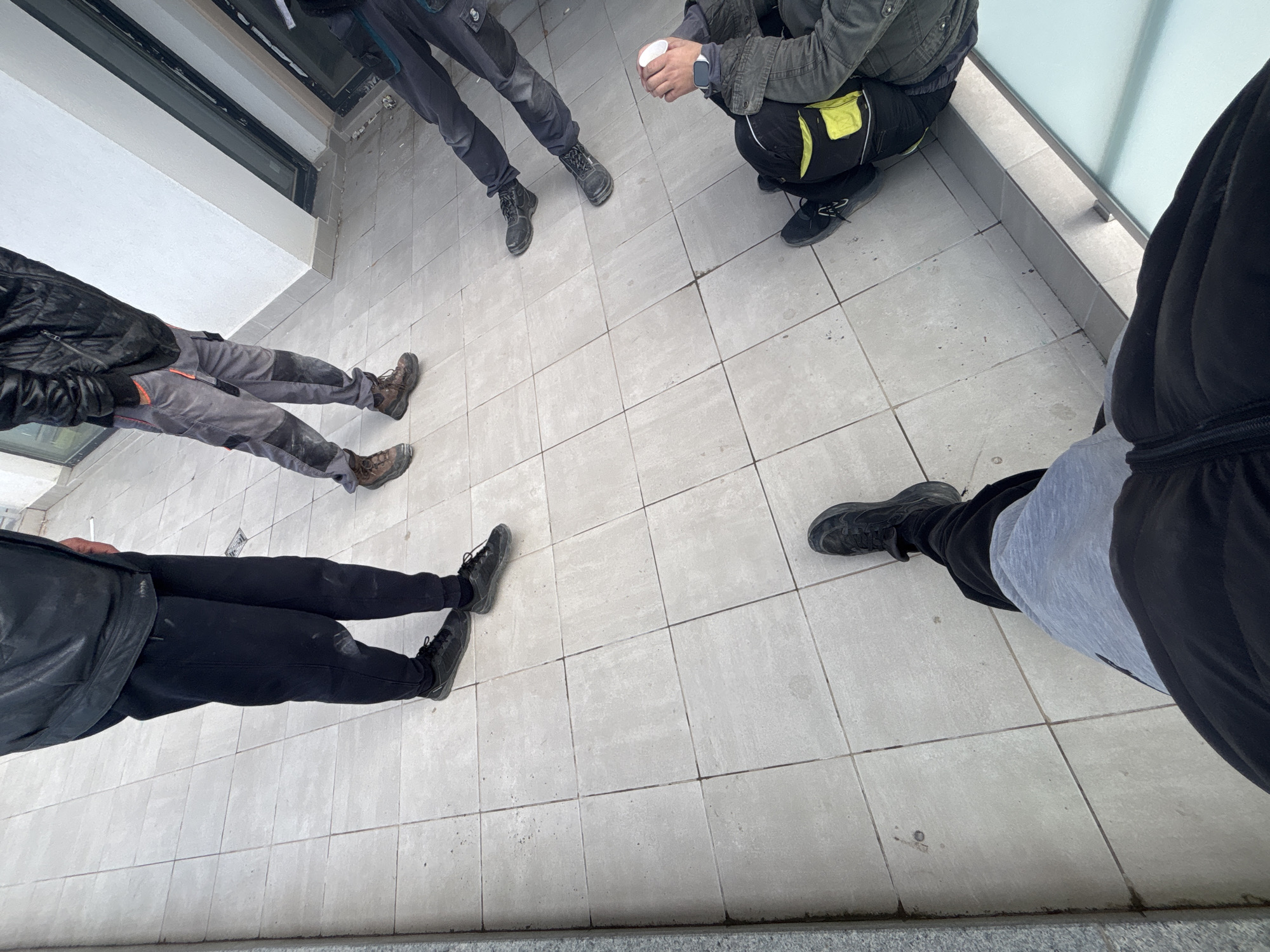 The image depicts a high-angle view of five individuals standing or crouching on a tiled surface. The location is likely an indoor or partially enclosed space, given the presence of what appears to be a window frame and a low wall or railing. The individuals are identifiable by their lower bodies and legs. The ground is a light gray tile. One individual is crouching, holding a white cup. The others are standing, and all are wearing work clothes and work boots. The environment is generally clean but unremarkable. The location within Plovdiv, Bulgaria, is indeterminable from this image alone. There is no obvious activity taking place aside from standing and crouching.