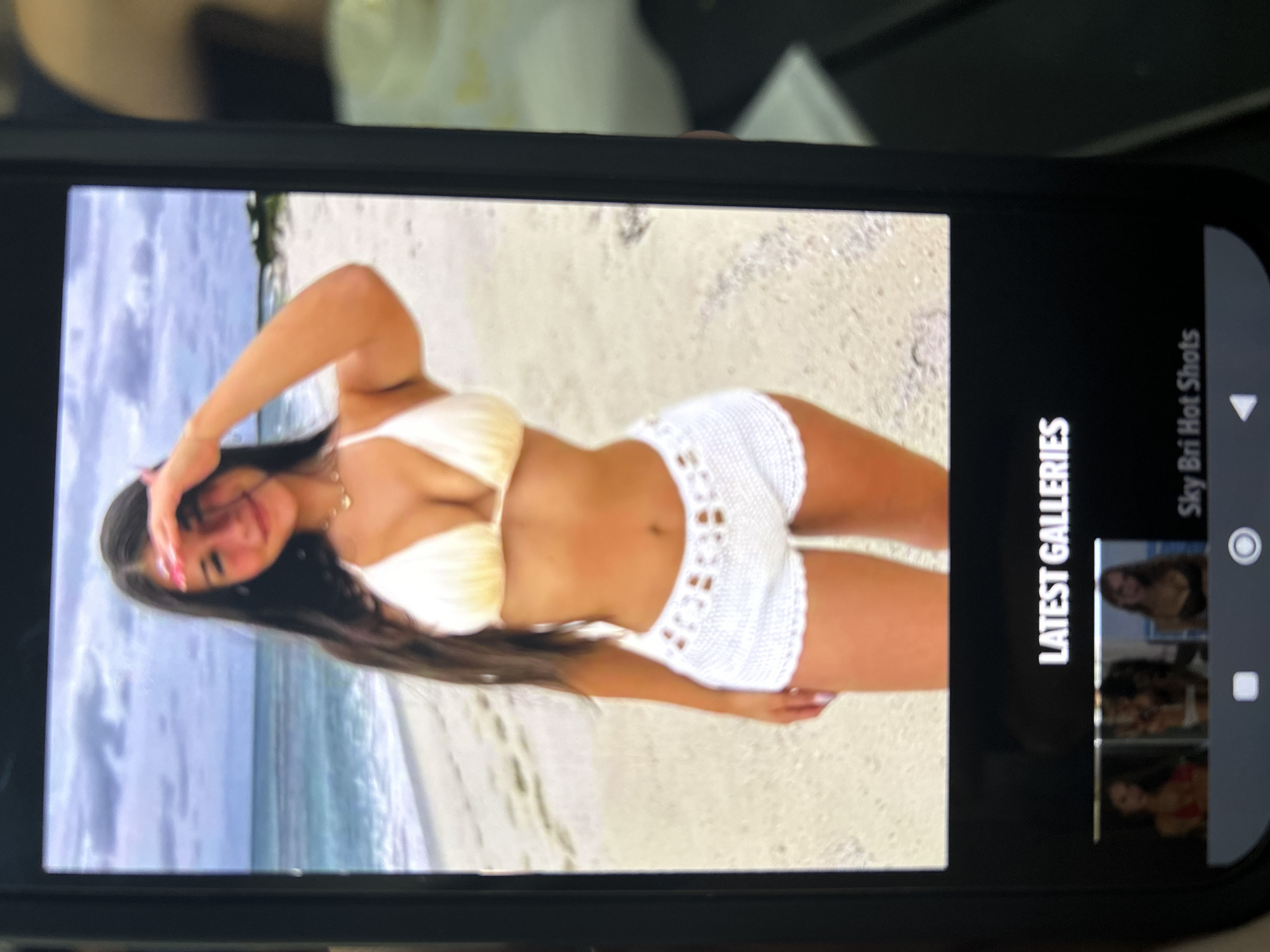 A photograph captures a dark-colored smartphone screen displaying a digital image and related content. The main image on the screen features a young woman standing on a beach. She has long, dark hair, smiles at the viewer, and uses her left hand to shield her eyes. She is wearing a light-colored, possibly white or cream, string bikini top and white crocheted shorts. Her fingernails are painted pink. The background of this image shows a sandy beach, blue ocean water with white waves, and a cloudy sky. Green foliage is visible on the distant shoreline to the right. Below the main image on the smartphone screen, the text "LATEST GALLERIES" is visible. Further down, a row of three blurred thumbnail images of women is displayed, followed by the partially visible text "Sky Bri Hot Shots." The smartphone's lower bezel and navigation icons are also visible. The scene depicted on the smartphone screen is located in Blagoevgrad, Bulgaria.