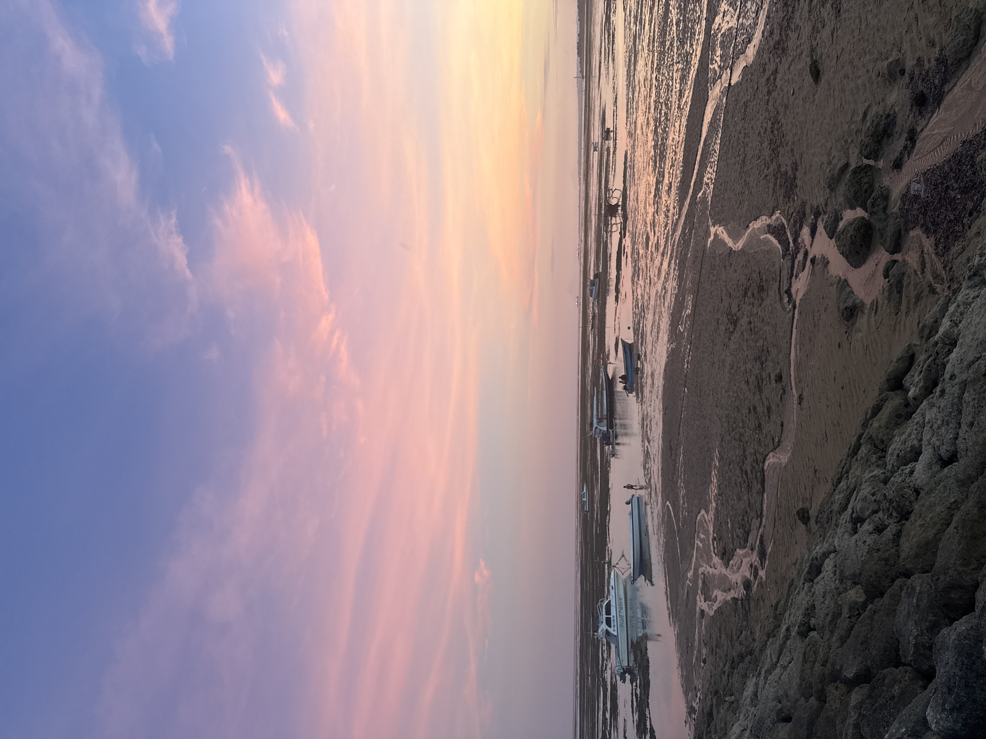 The image depicts a coastal scene in Denpasar, Indonesia, during a vibrant sunset. The sky blends purple, pink, and yellow hues. The tide is low, exposing a muddy shoreline with meandering rivulets. Several boats rest on the tidal flats. A rocky embankment forms the foreground on the right.