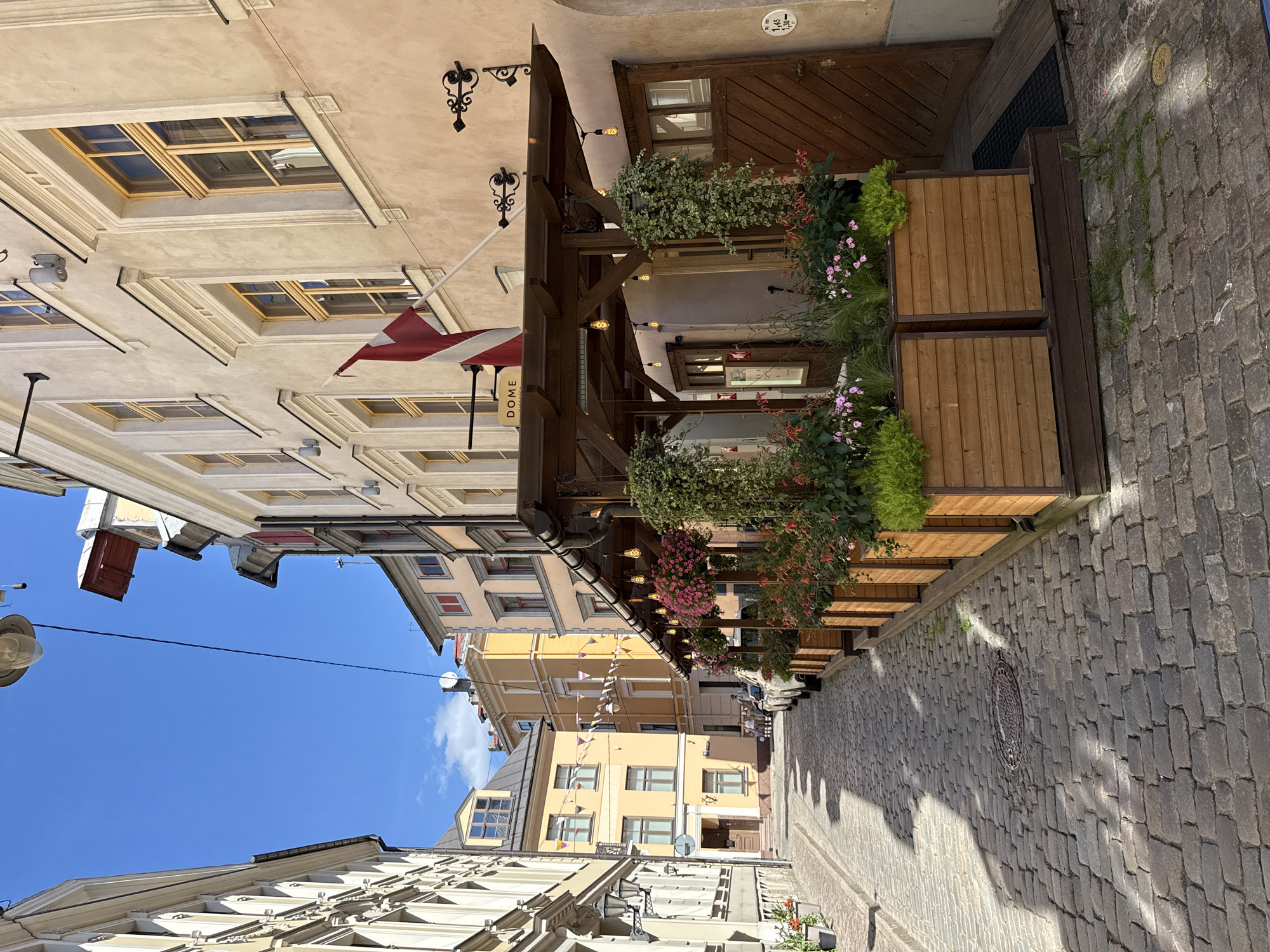 The image depicts a narrow, cobblestone street in the Old Town of Riga, Latvia, on a sunny day. The street is flanked by historic buildings with light-colored facades and yellow-framed windows. On the right side of the street, a restaurant or cafe has an outdoor seating area with wooden planters filled with vibrant pink and green flowers. A wooden awning covers the seating area, which is illuminated by decorative string lights. A Latvian flag flies above the awning. The cobblestone street is partially shaded by the buildings. In the background, other buildings with colorful facades and a clear blue sky with a few wispy clouds are visible. A few parked cars and a small group of people can be seen further down the street. The scene conveys a charming and picturesque atmosphere of a European city.
