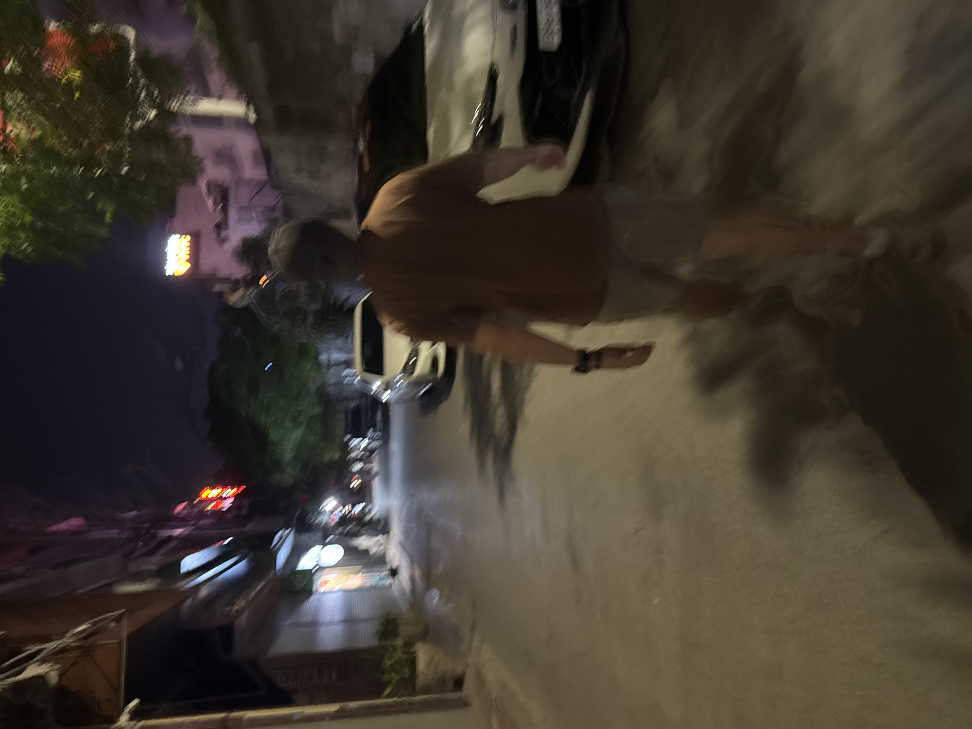 The image depicts a person walking down a street at night in Hanoi, Vietnam. The street appears to be narrow and lined with buildings and parked cars. There are some neon signs visible on the buildings, indicating the presence of businesses. The lighting is dim, with some artificial lights illuminating the scene. The person in the foreground is walking away from the camera, wearing a brown t-shirt and shorts. The overall atmosphere is that of a typical urban street at night.