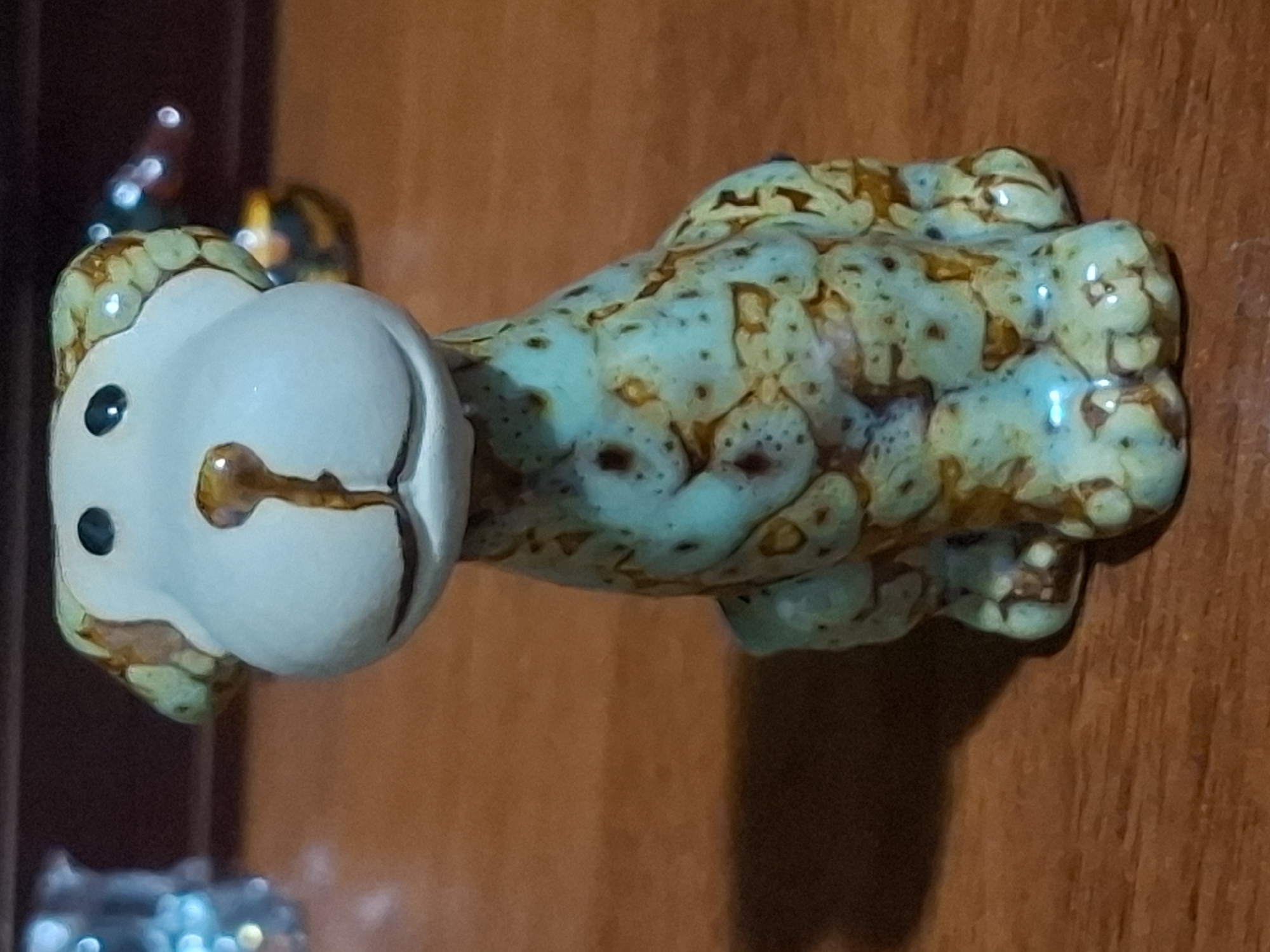 A small, ceramic figurine of a dog is positioned on a wooden surface. The figurine has a light blue face with black eyes and a brown nose. Its body is decorated with a mottled pattern of turquoise and brown. The figurine is sitting and facing forward. The background is blurred but appears to be a dark shelf or wall with some out-of-focus lights. The wooden surface suggests an indoor setting, possibly a home or display area. There is no discernible location in the city of Almaty, Kazakhstan, mentioned or implied in the image.