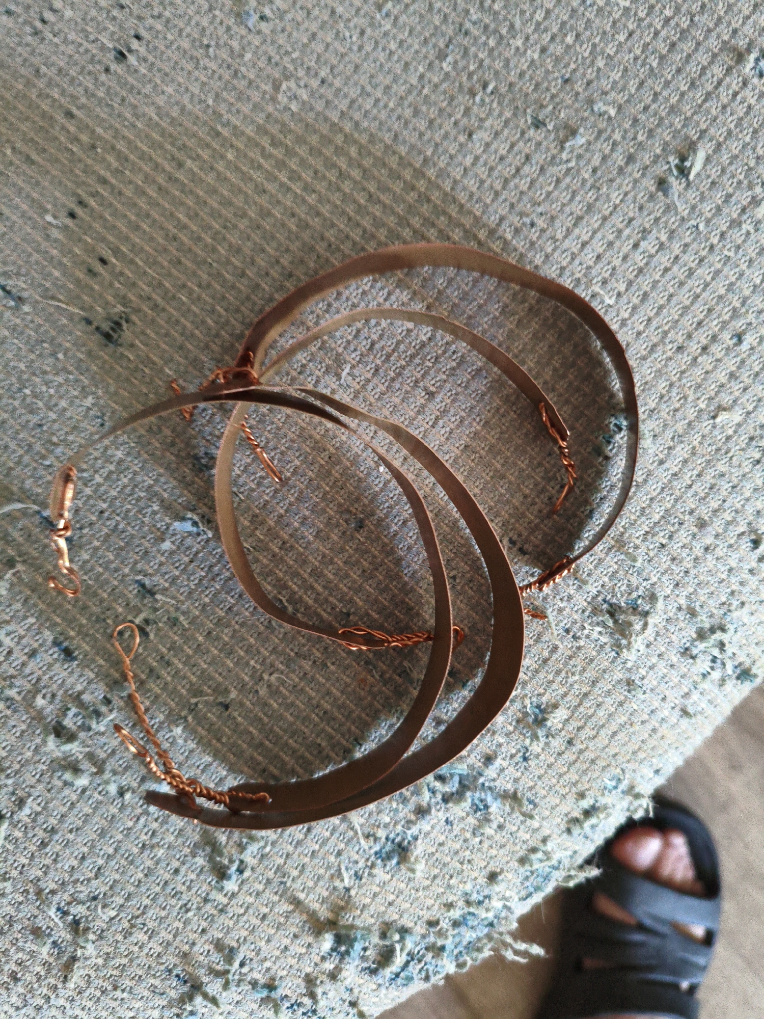 This media file features a close-up, overhead shot of several flat, metallic-looking, crescent-shaped objects, possibly jewelry components or craft supplies, lying on a textured, light blue-grey fabric surface. The objects appear to be made of copper or a similar reddish-brown metal and are arranged in a circular, overlapping pattern. Small twists of finer copper wire are visible, securing or decorating some of the larger pieces. In the lower right corner, a person's bare foot wearing a black sandal is partially visible, suggesting a casual, indoor setting. The fabric surface has a woven texture with some darker specks and small patches of blue fiber, hinting at a worn or utilitarian textile, perhaps upholstery or a rug. The lighting is diffused, and there are no strong shadows, indicating it might be an overcast day or the shot was taken indoors with ambient light. No text is visible in the image. The scene is static, with the objects laid out and seemingly awaiting further assembly or use. The overall impression is one of a craft or DIY project in progress.