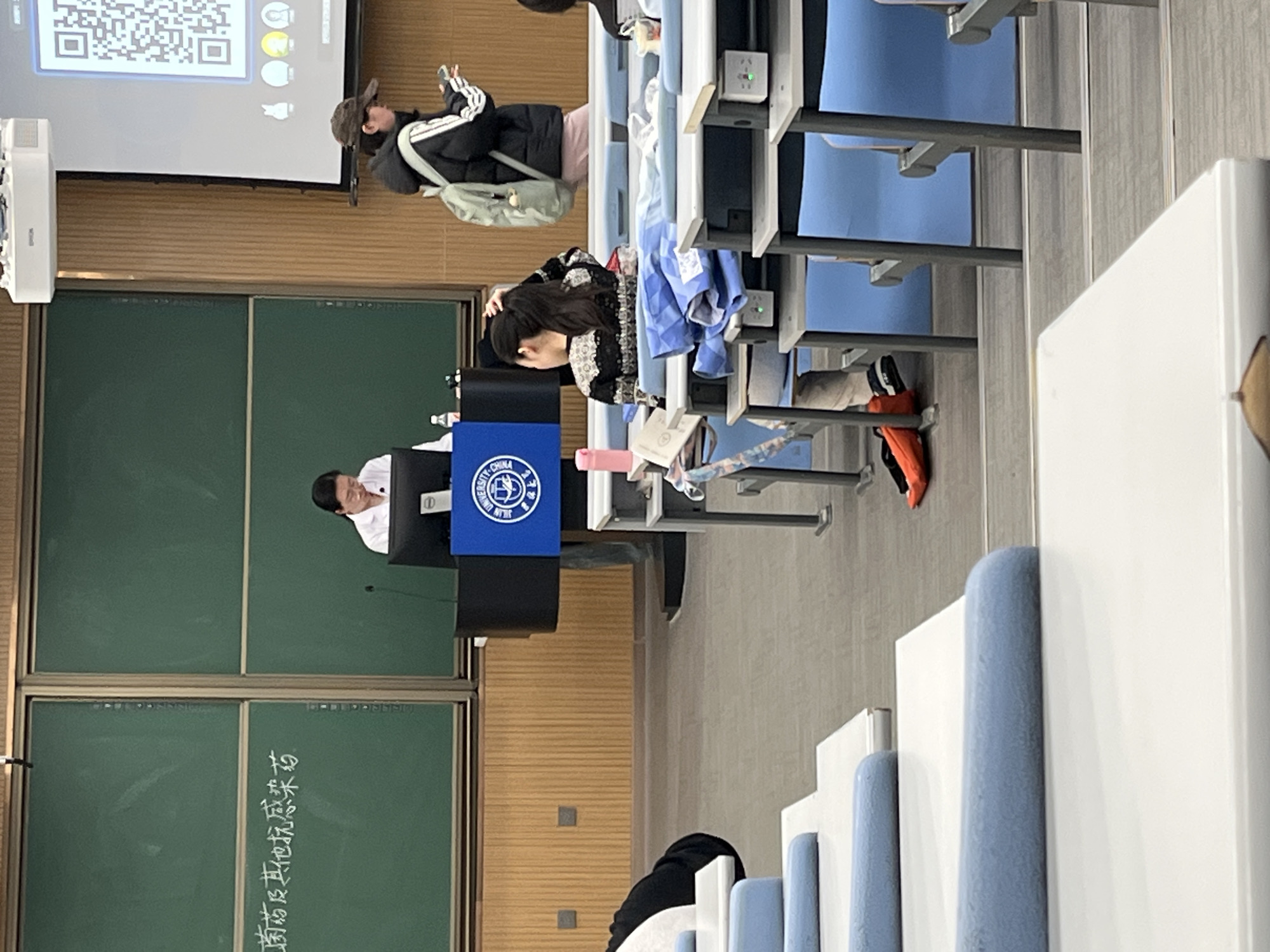 The image depicts a classroom scene, likely in a university setting within Changchun City, China, given the presence of signage. A professor stands at a lectern facing the students, and a projection screen displays a QR code. Seated students occupy the rows of desks. The chalkboard behind the professor has writing on it in Chinese. The room appears to be well-lit and orderly. The classroom is equipped with standard lecture hall furniture, including desks with integrated seating, individual power sockets, and writing surfaces.
