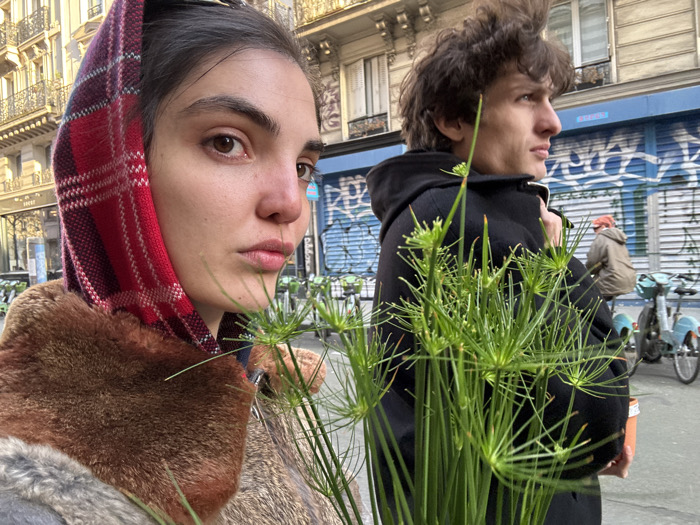 The image captures a street scene in Paris, France, featuring two individuals in the foreground. A young adult female on the left, wearing a red and purple plaid head covering and a brown fur-collared coat, looks directly at the camera. To her right, a young adult male with dark, curly hair, dressed in a black hooded coat, directs his gaze to the right of the frame. A cluster of green umbrella papyrus plants is held between them, prominent in the lower foreground. In the mid-ground, a row of blue Vélib' public bicycles is parked along the curb. A third, partially obscured individual wearing a beige jacket and an orange cap is seated on one of these bicycles. The background on the left displays a classic Haussmannian building facade. On the right, a building with a blue storefront, marked with graffiti, is visible.