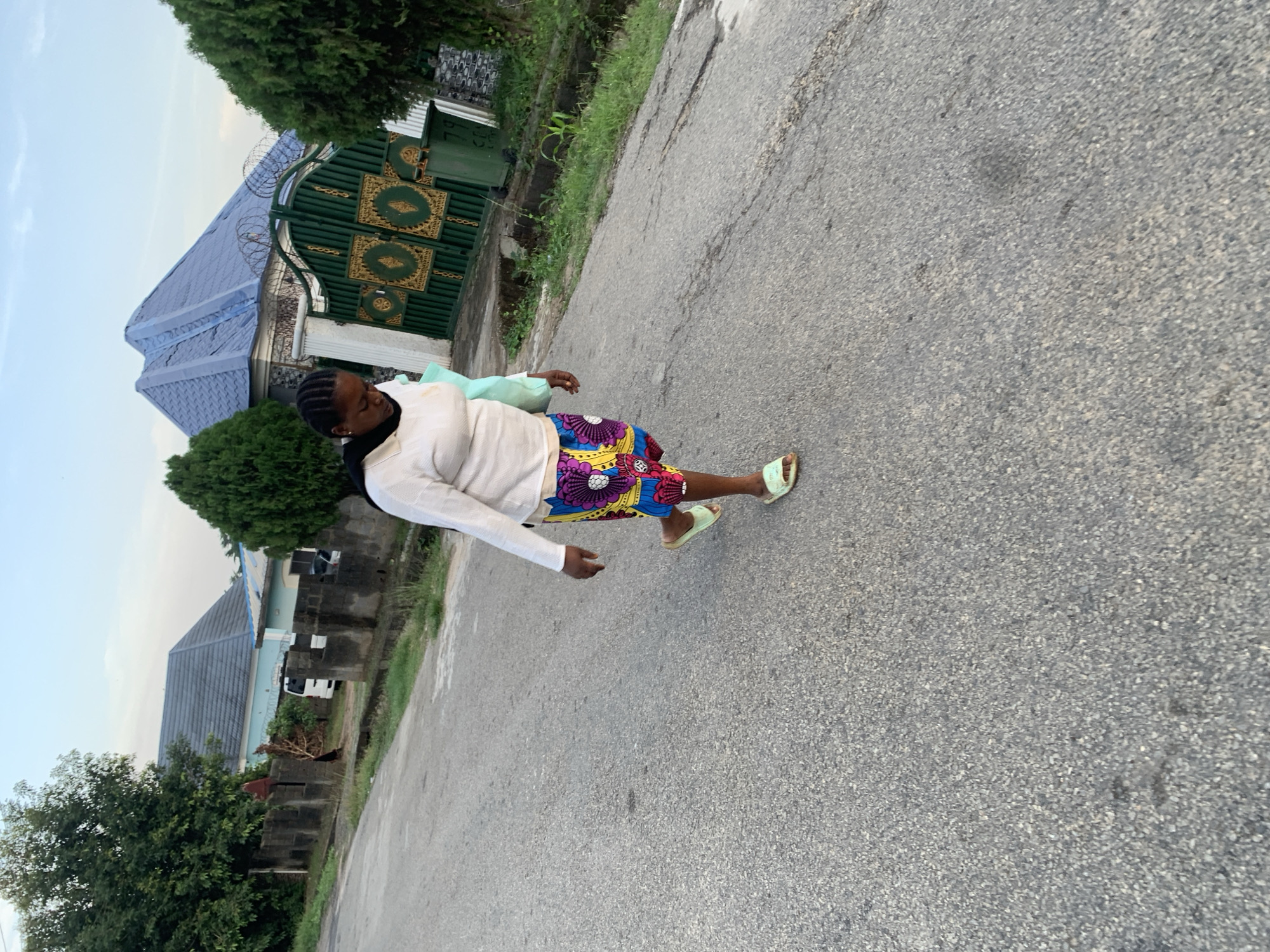 A woman is standing on an asphalt road in what appears to be a residential area in Abuja, Nigeria. She is wearing a long-sleeved white top, a colorful patterned skirt, and light green sandals. The road is cracked and shows signs of wear.
To the left of the woman, there are houses with blue roofs and green gates visible. There are also trees and shrubs. The sky is light, suggesting it is either early morning or late afternoon. The image is taken at a slightly tilted angle.