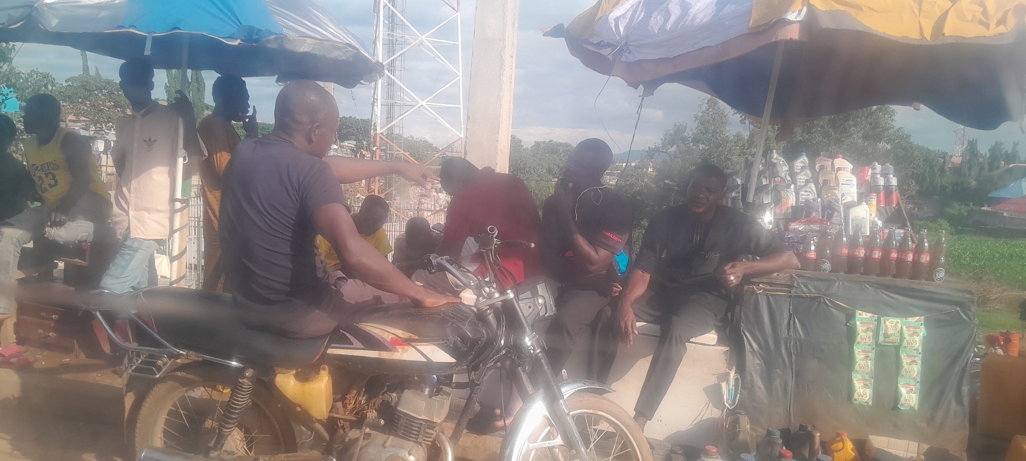 In Nyanya, Nigeria, a street vendor is seen operating under an umbrella. A motorcycle is parked in the foreground. The vendor displays various items for sale, including bottles of Coca-Cola, several cans of lubricant oil, and other assorted goods. A group of people are gathered around the stall. One man, wearing a dark shirt, is seated and appears to be conversing. Another man, in a maroon shirt, is partially visible. Several other individuals are also present, some standing and some seated. The setting appears to be an outdoor market area, with a tall structure resembling a cell tower in the background. The overall scene depicts a typical roadside market environment.