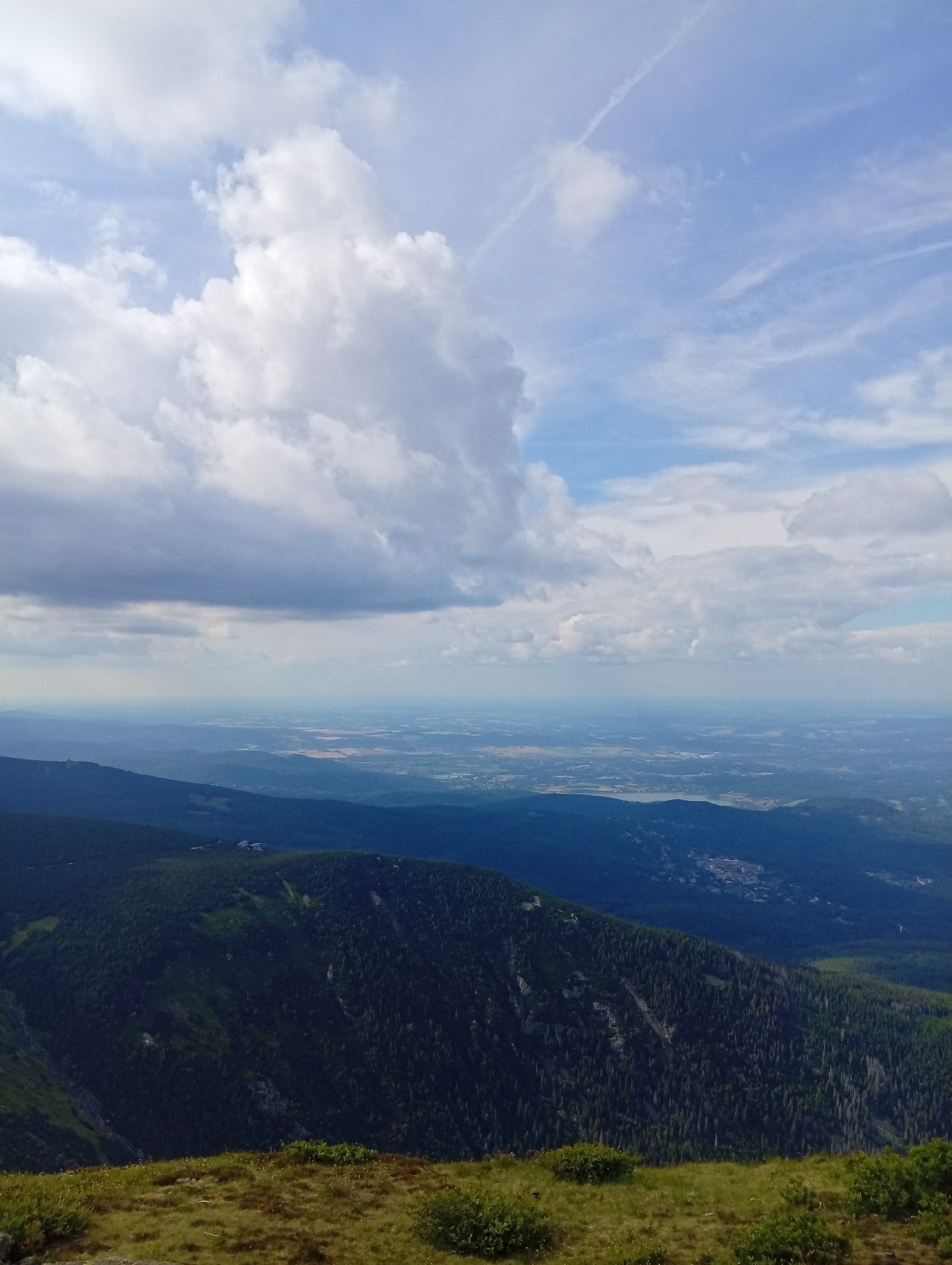 This is a high-angle, full shot of a mountainous landscape in Malá Úpa, Czechia. The foreground is a grassy slope with small yellow flowers and green bushes. The middle ground is dominated by a dark, forested mountain range with a visible trail. In the background, layers of mountains recede into a hazy, distant landscape with a visible body of water and a town. The sky is filled with dramatic clouds, ranging from dark storm clouds to bright white cumulus clouds against a pale blue sky. There are no people or animals visible in the image.