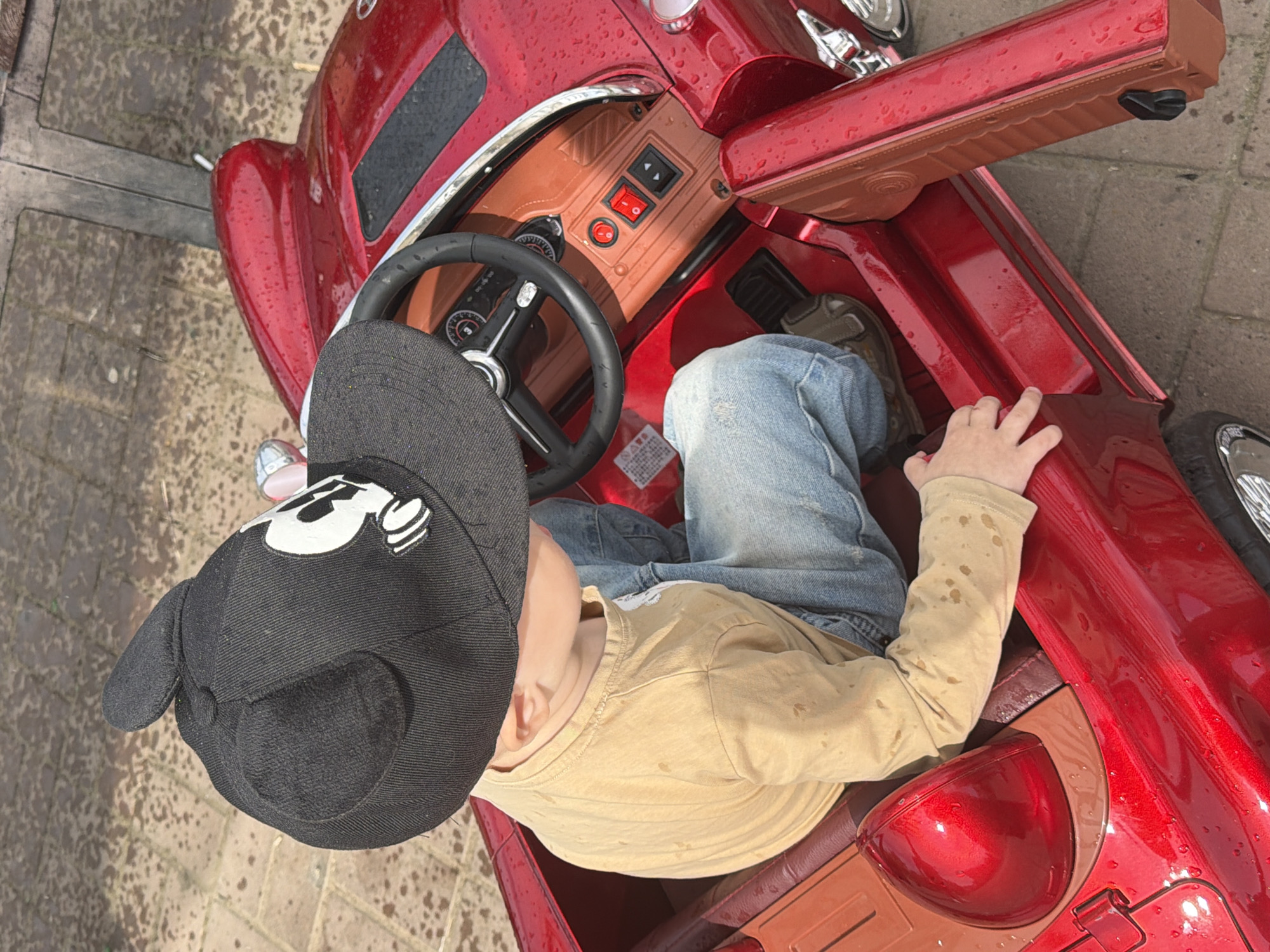 A young child is seated in a red toy car. The child wears a black baseball cap with mouse ears and a Mickey Mouse graphic, a tan long-sleeved shirt, and blue jeans. The toy car has a brown interior, a steering wheel, and dashboard controls. The car is parked outdoors on a paved surface, possibly a sidewalk or driveway. The lighting suggests it is daytime. There are water droplets on the car's surface, indicating recent rain or mist. The environment appears to be an urban setting. The child's hands are visible, one resting on the edge of the car door and the other near the car's interior.