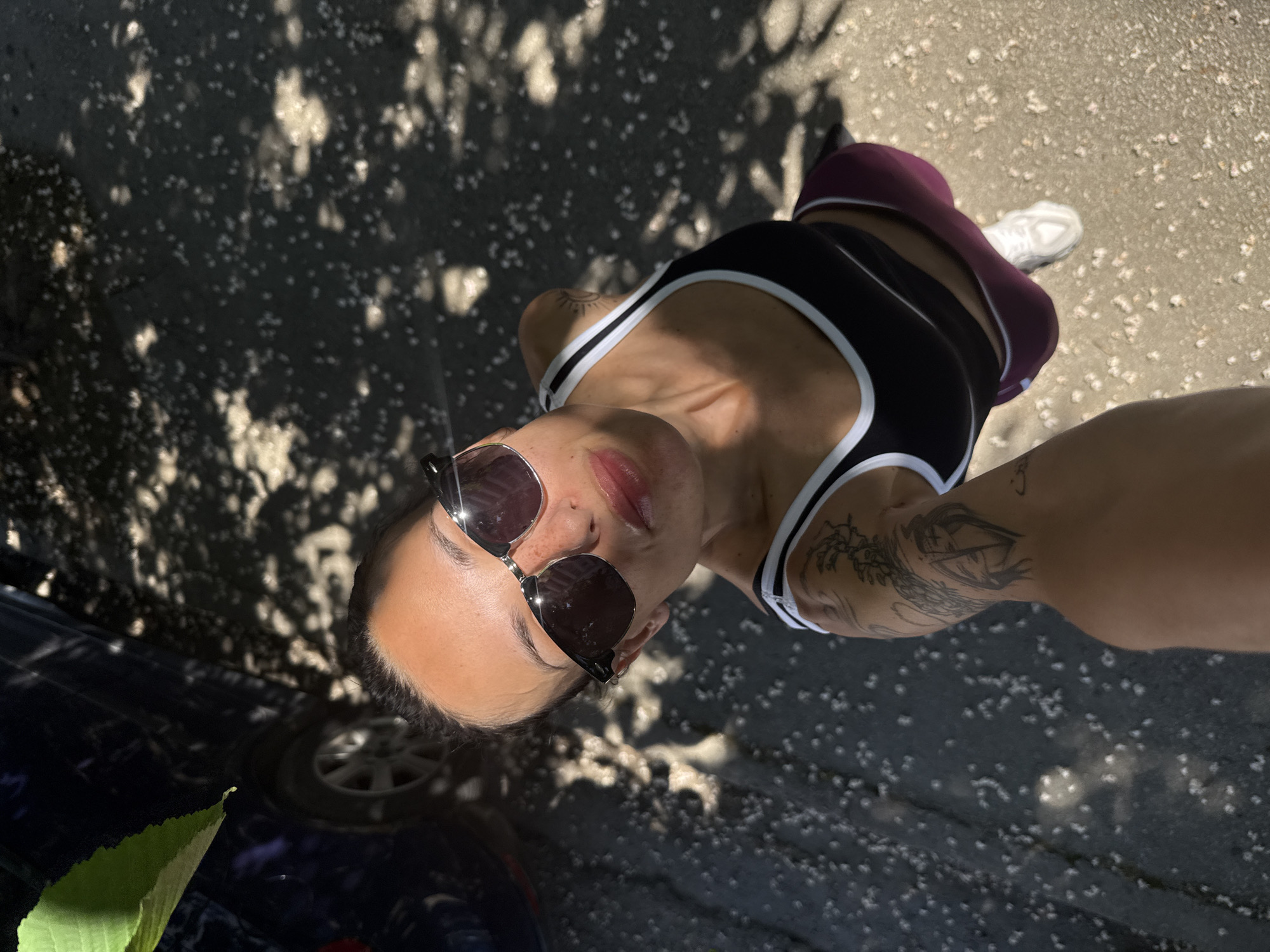 A selfie taken outdoors in the city of Pleven, Bulgaria shows a light-skinned individual wearing sunglasses, a black tank top with white trim, maroon leggings, and white shoes. The subject is standing on what appears to be a paved surface covered with scattered white petals or specks of light. Part of a dark blue car is visible in the lower left corner, with a green leaf near its front wheel. The scene is dappled with sunlight filtering through trees, creating a pattern of shadows and light on the ground. The perspective is from below, angled upwards towards the subject.