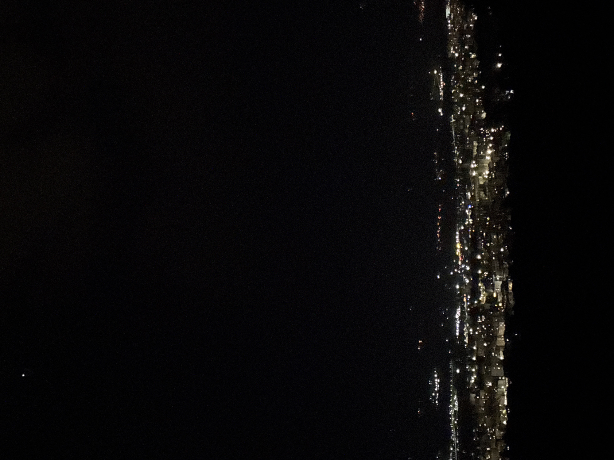 The image shows a nighttime cityscape. Bright lights are clustered along the right side, suggestive of buildings, possibly near a body of water. The left side is predominantly dark, indicating open space or areas with minimal lighting. The location is indeterminate due to the limited visual information. There is no evidence to support that this picture shows the city of Marulevo, Bulgaria. There is no discernible information to identify people, subjects, objects, actions, or interactions.
