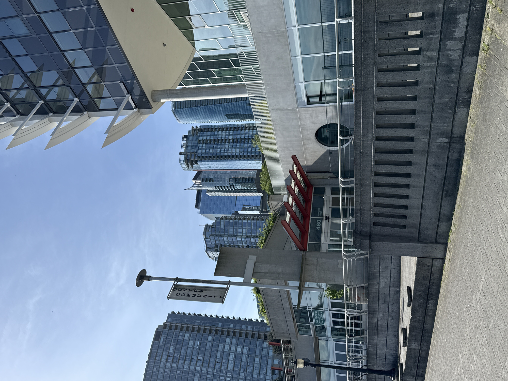 The image shows the Coal Harbour neighborhood in Vancouver, Canada, with a view of several buildings and landmarks. The architectural style is modern, featuring glass and concrete structures. In the foreground, there is a sign pointing to the Coal Harbour Community Centre. The surrounding buildings include residential towers and commercial spaces. The sky is blue with some light clouds, indicating a sunny day. The scene captures the urban environment and design aesthetic typical of Vancouver's waterfront.