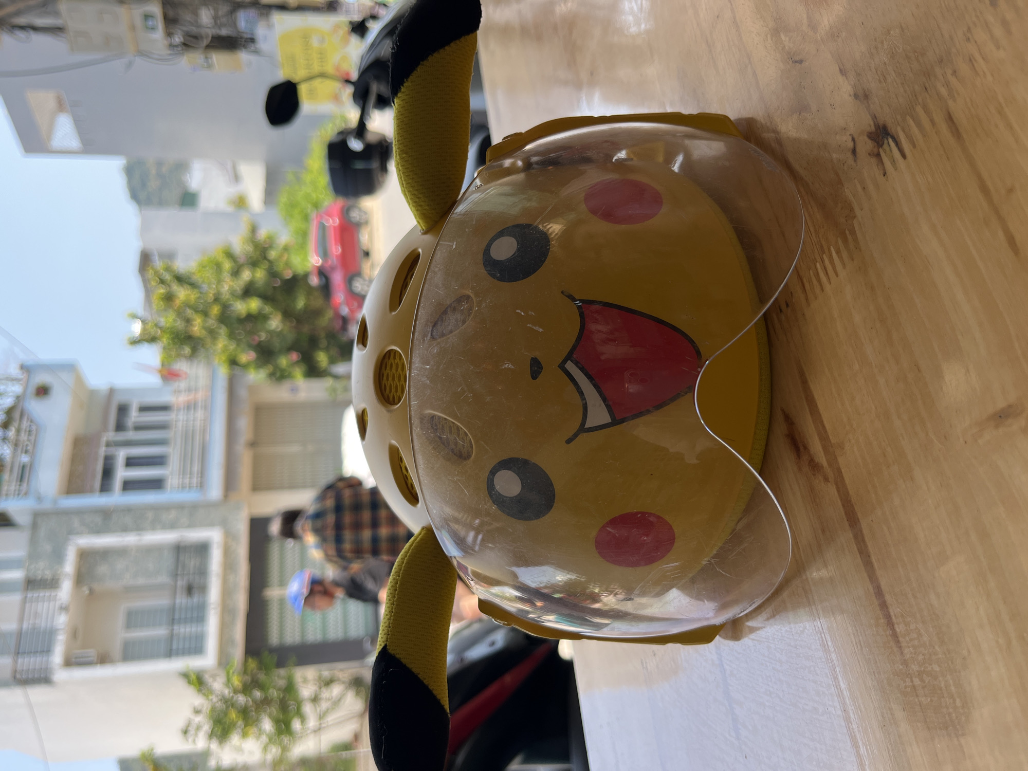 The image shows a yellow children's bicycle helmet designed to look like Pikachu, resting on a wooden surface. The helmet has black and yellow ears, red cheeks, and a large, open-mouthed smile. A clear plastic visor is attached. Outside, a street scene is visible, including buildings, trees, a red car, and a motorcycle. A person wearing a plaid shirt and another wearing a blue helmet are also visible in the background. The location appears to be an urban area in Unknown.