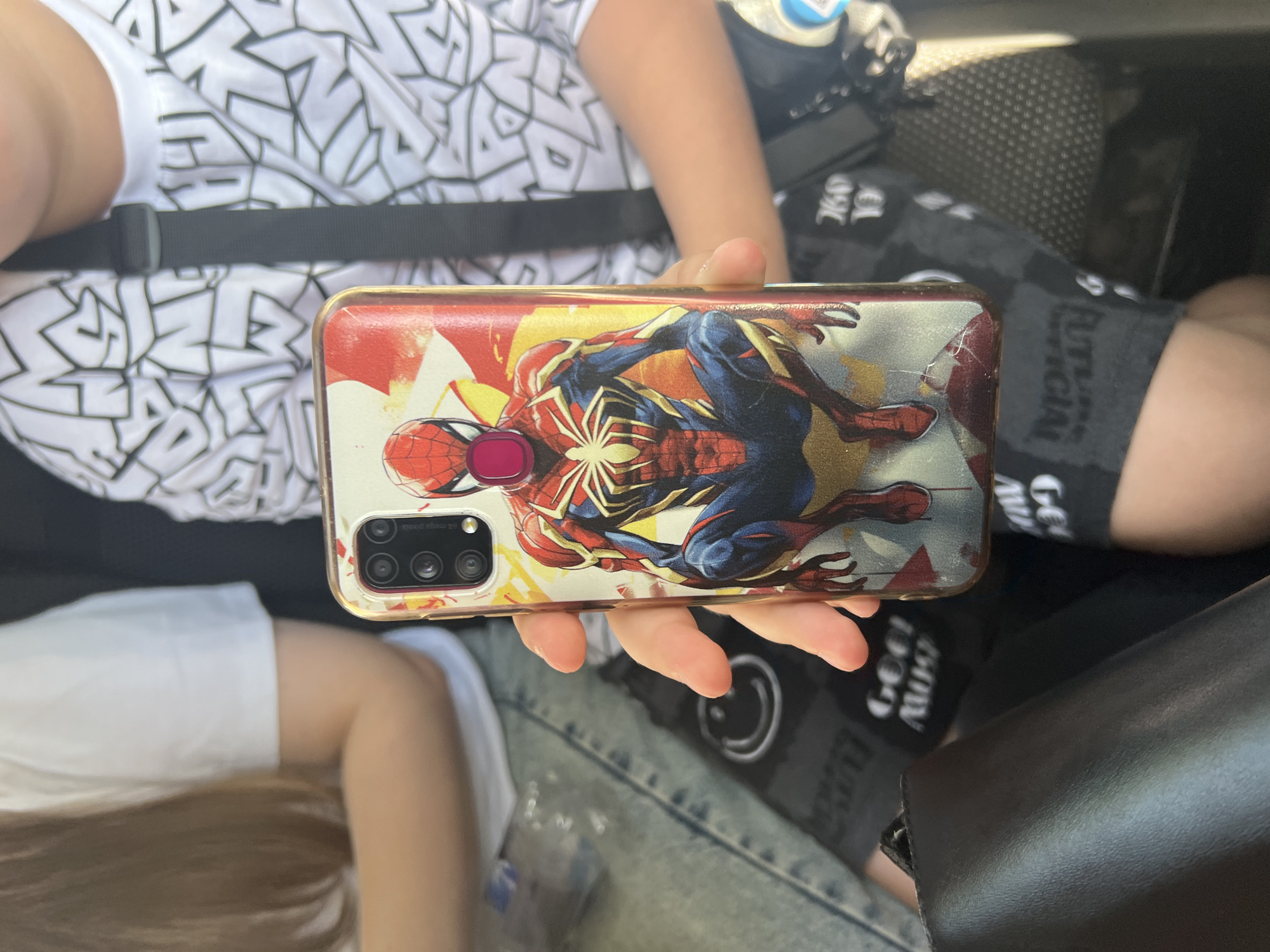In Ulyanovsk, Russia, a person is holding a mobile phone with a Spider-Man-themed case. The case features a vibrant depiction of the superhero in a dynamic pose against a backdrop of reds and yellows. The person's hands are visible, one on the side of the phone, and the other is obscured. The background shows parts of two other individuals, one wearing a white t-shirt with long brown hair, and another wearing a white t-shirt with a black and white graphic pattern. The overall environment appears to be indoors, possibly a vehicle, given the dark seats and straps visible.