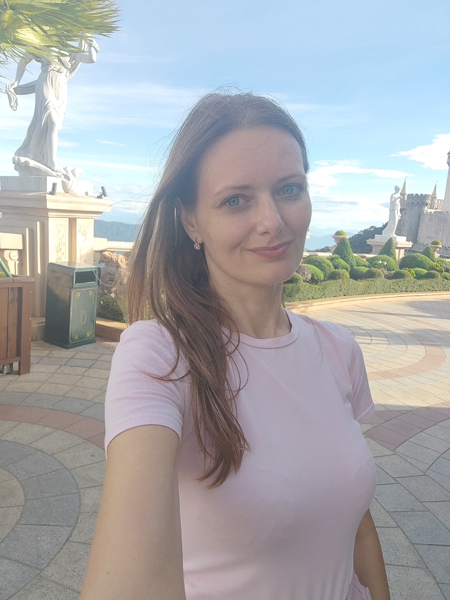 A woman with long brown hair and blue eyes, wearing a light pink t-shirt, takes a selfie outdoors. She is standing on a paved area with circular patterns, with a white statue of a woman holding a drapery and a palm frond visible on the left. In the background, there are manicured bushes, a castle-like structure with spires, and mountains in the distance. The sky is blue with scattered clouds. A green trash bin with decorative patterns is situated to the left of the woman. The overall scene appears to be within a tourist attraction in Đà Nẵng, Vietnam, potentially the Ba Na Hills resort, known for its European-inspired architecture and landscapes.