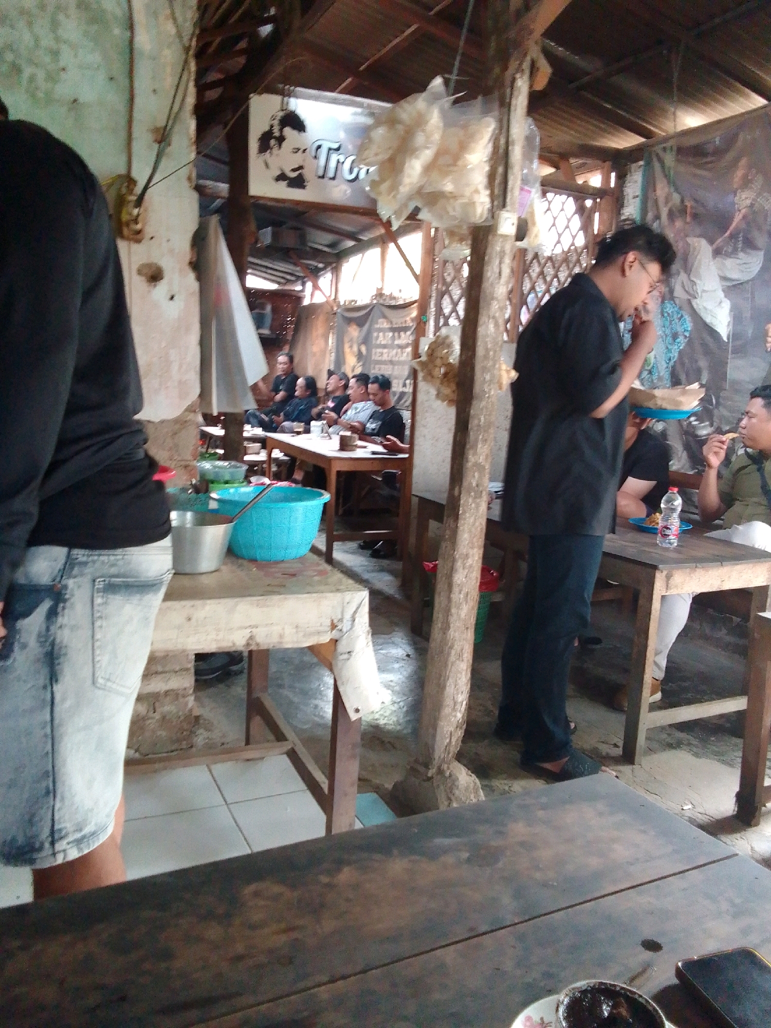 A group of men are seated at tables in a rustic outdoor eatery. The tables are made of rough wood, some covered with white cloths. In the foreground, a man in a black shirt and blue jeans stands facing away from the camera, his back and left side visible. In the middle ground, to the right, a man in a black shirt is eating from a plate. Further back, several men are seated at a table, some of whom appear to be engaged in conversation. A blue plastic basket and a metal pot are on a table in the foreground. The eatery has a wooden roof supported by posts. Bags of snacks hang from the ceiling. A sign with a face and the word "Trom" is visible overhead. The scene appears to be set in Trenggalek, Indonesia, given the presence of the "Trom" sign, which is likely an abbreviation for Trenggalek.