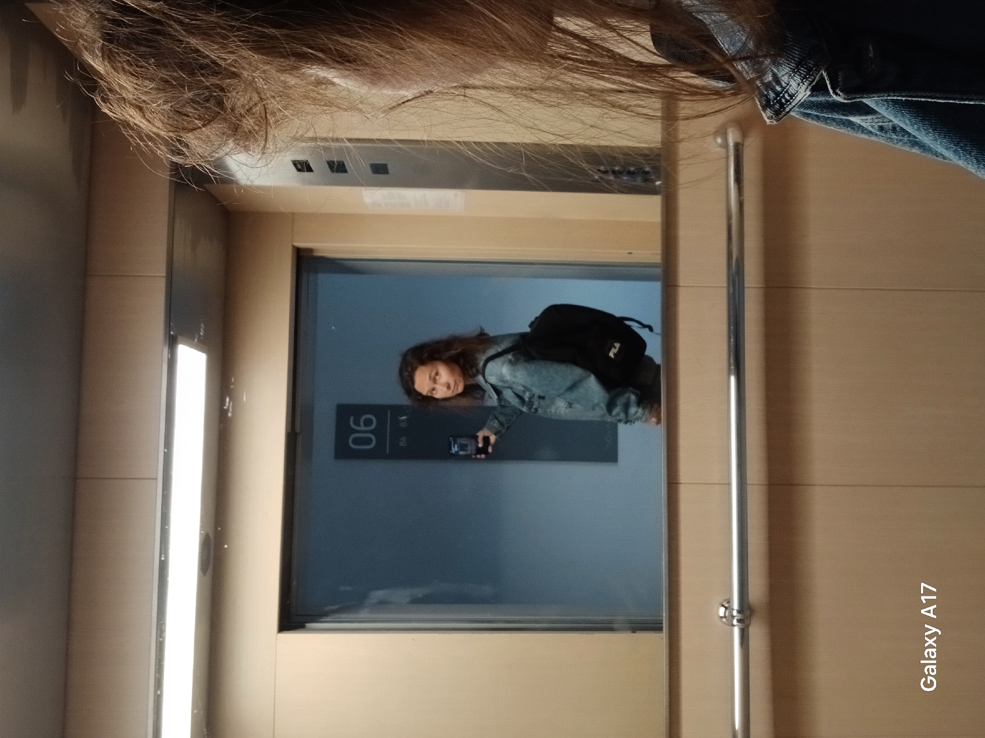 The image, rotated 90 degrees clockwise, captures a scene from within an elevator in Yekaterinburg, Russia. In the foreground, the top of a person's head with brown hair and a blue denim jacket sleeve are visible, indicating the perspective of the photographer inside the elevator. Through a glass panel, another person, likely female, is seen standing outside the elevator in what appears to be a hallway or landing.
This individual has long, dark, wavy hair and is wearing a light blue denim jacket over a dark top. She carries a black backpack with a white "FILA" logo on her back. In her right hand, she holds a dark-colored smartphone, looking directly towards the camera with a neutral to slightly contemplative expression. Behind her, on a dark panel, the number "06" is clearly visible, suggesting the elevator is at the sixth floor. The elevator's interior features light-colored, possibly wooden or laminate, panels and a vertical chrome railing on the right side. A bright vertical light strip illuminates the left side of the elevator entrance.
The bottom right corner of the image, when rotated correctly, displays a watermark that reads "Galaxy A17," indicating the device used to capture the photograph.