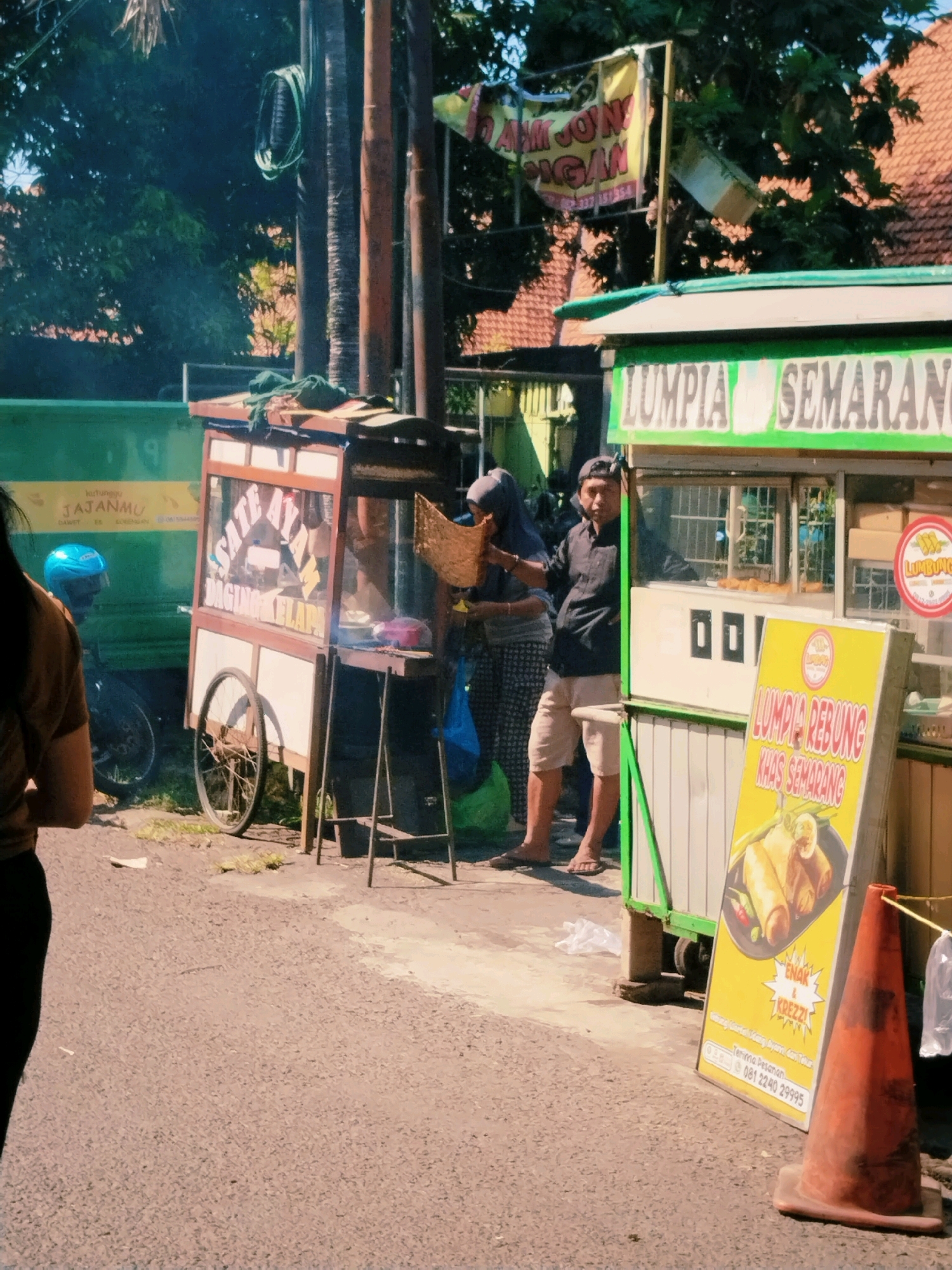 In Surabaya, Indonesia, on a sunny day, a street scene unfolds with vendors and pedestrians. A green truck with yellow text displaying "KUTUNGGU JAJANMU" is parked on the left. Next to it, a food cart with wooden frames and glass panels, adorned with text suggesting "Sate Ayam Daging Kelapa", is positioned. A man wearing a black shirt and beige shorts stands behind this cart, interacting with a woman in a gray headscarf and patterned skirt who is holding a brown paper bag.
Further to the right, a food stall with a green and white awning advertises "LUMPIA SEMARANG" in large letters. A yellow sign in front of this stall features an image of spring rolls and text including "LUMPIA REBUNG KHAS SEMARANG," along with a phone number. An orange traffic cone stands in the foreground, and various plastic bags are visible on the ground. The ground is paved with asphalt, and vegetation is present in the background, suggesting a bustling urban environment. A blue motorcycle helmet is visible on the left side of the image.