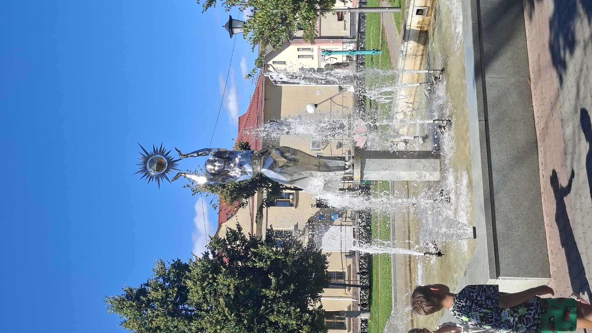 A full shot captured on a sunny day shows a gleaming silver sculpture of a woman holding a sun above her head, centrally located in a public fountain in Pastavy, Belarus. The sculpture, made of reflective metal, stands on a grey pedestal surrounded by multiple streams of water shooting upwards, creating a dynamic spray. In the foreground, a young boy wearing a dark t-shirt with a colorful graphic print and green shorts is partially visible from the side, looking towards the fountain. His shadow is cast on the patterned pavement. Behind the fountain and sculpture, a yellow building with a red-tiled roof is visible, along with lush green trees and a clear blue sky. A lamppost stands to the right of the building. The environment appears to be a well-maintained public space, likely a town square or park.