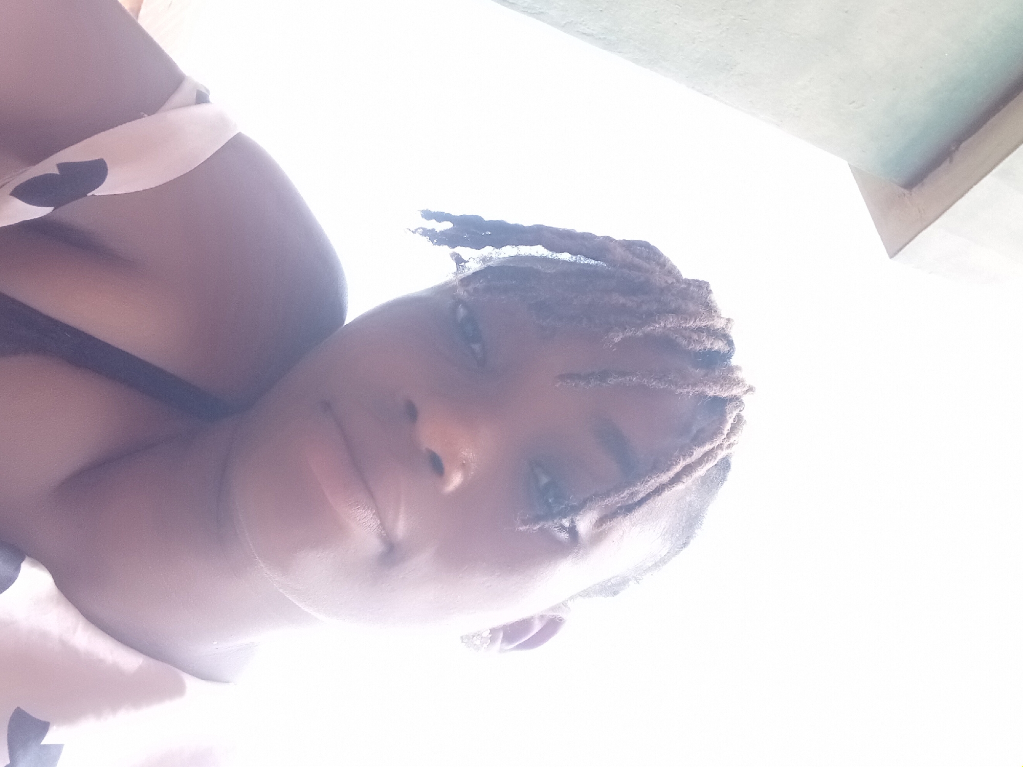A close-up, low-angle selfie shows a woman with braided hair, light skin, and brown eyes. She wears a black bra. Her skin has a light shine. She wears a white patterned top. The background is washed out by the light. The location is Shagamu, Nigeria.