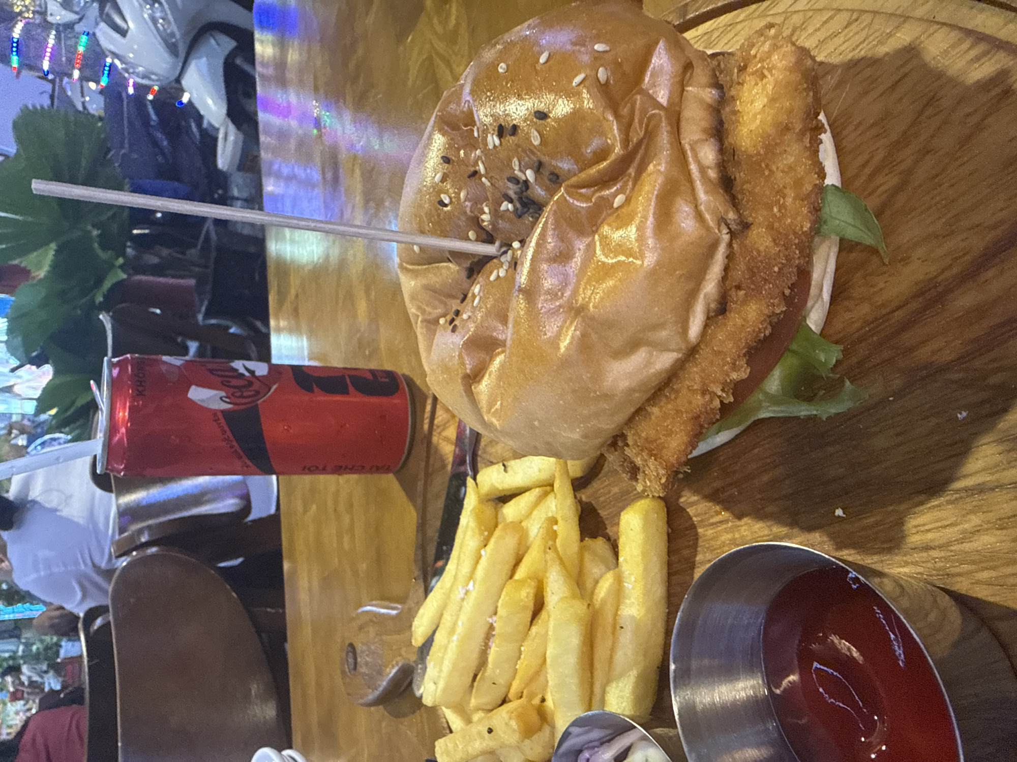 The image shows a wooden table with a hamburger, french fries, and a can of Coca-Cola. The hamburger is large and topped with sesame seeds, with a skewer inserted to hold it together. A side of french fries and a small metal container of ketchup accompany the burger. The Coca-Cola can is red and partially visible next to the food. In the background, a blurred cityscape or outdoor dining area is visible with a partially visible scooter. The location appears to be in a city, but the specific city and country are unknown.