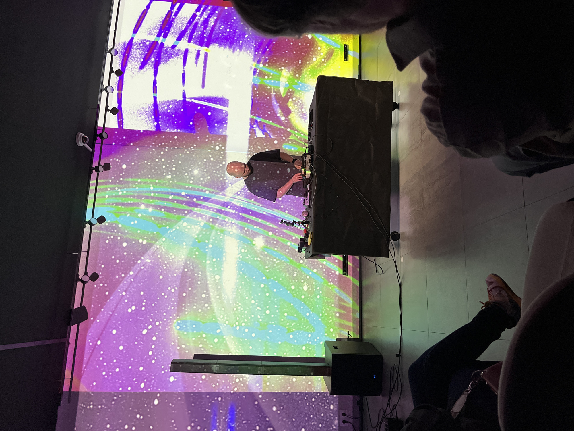 The image shows a live music performance in a dark indoor space with a large screen displaying colorful, abstract, possibly psychedelic, visuals. A bald male DJ in a dark shirt is positioned behind a setup of electronic music equipment, including synthesizers, mixers, and possibly other audio processing units. The DJ appears to be actively performing or adjusting the equipment.
In the foreground, parts of people are visible, suggesting the perspective of an audience member. A foot wearing a tan sneaker and dark pants is noticeable on the right. Other attendees are partially visible, indicating the presence of a crowd watching the performance.
The background shows a black wall with multiple round lights positioned along it.
The city and location are not discernible from the provided image.
