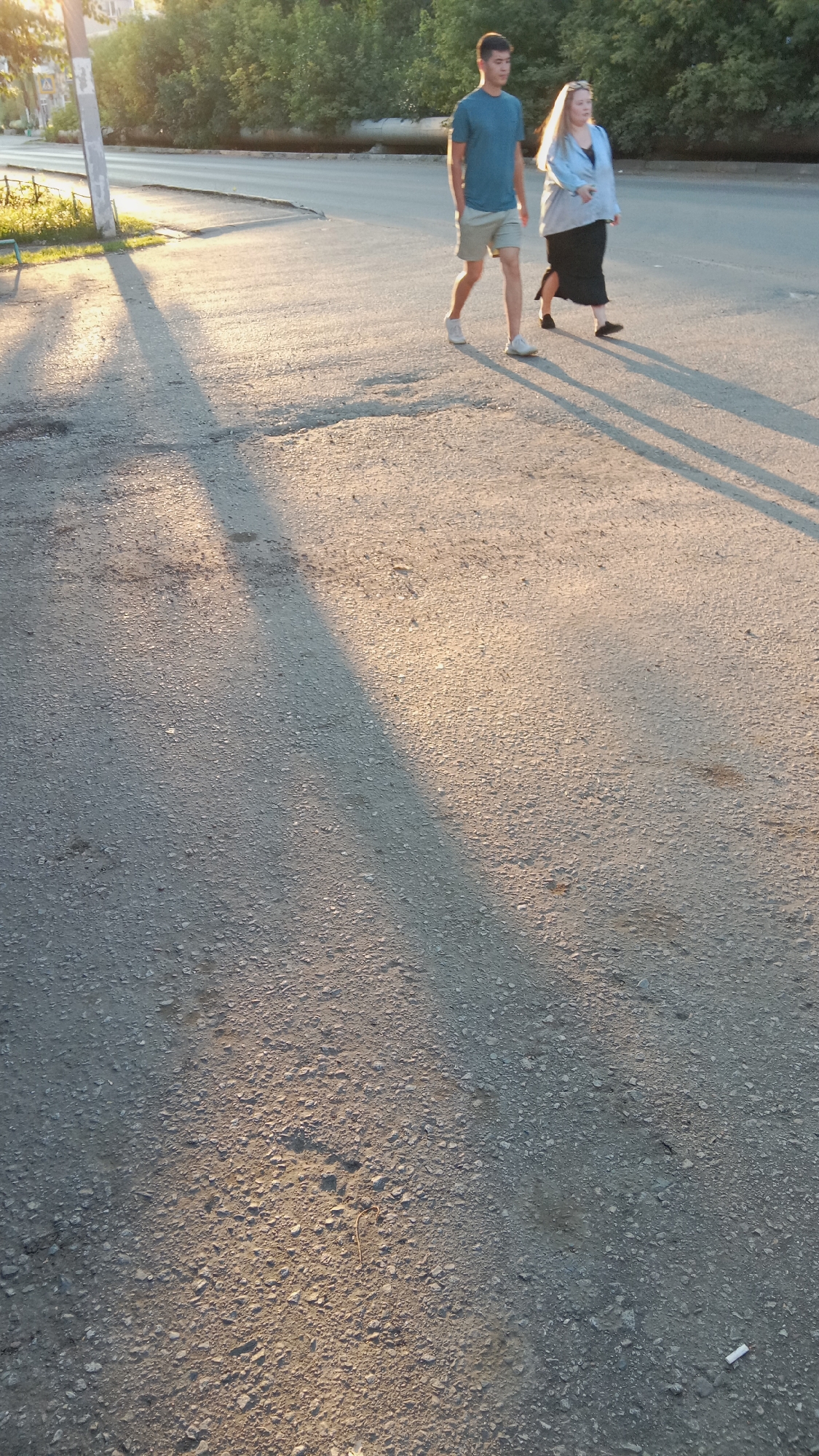 Two individuals, a male and a female, are walking on an asphalt surface in Petropavl, Kazakhstan. The male is wearing a blue t-shirt and light-colored shorts with white sneakers, while the female is wearing a light blue jacket over a darker garment and a dark skirt, with black shoes. They are walking away from the viewer and are casting long shadows on the ground due to the low angle of the sun. The environment appears to be an urban or suburban street with trees and a building visible in the background. The lighting suggests it is either early morning or late afternoon.