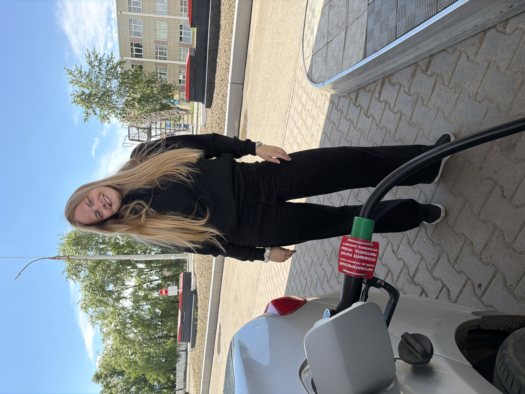 A young woman with long blonde hair stands at a gas station in Chaikovsky, Russia. She is dressed in a black tracksuit. A silver car is visible with a fuel nozzle inserted into its gas tank. The gas pump displays text in Russian. The surrounding environment includes a building, trees, a blue sky, and paved ground.