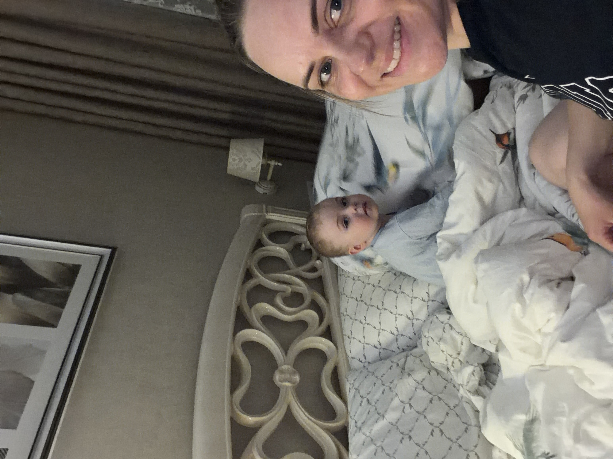 A woman and a baby are in bed. The woman is smiling at the camera. The baby is lying on its side, looking up. The headboard of the bed is ornate and off-white. There is a lamp on a nightstand and a framed picture on the wall. The bedclothes appear to be patterned with a grey design. The location is not discernible from this image.