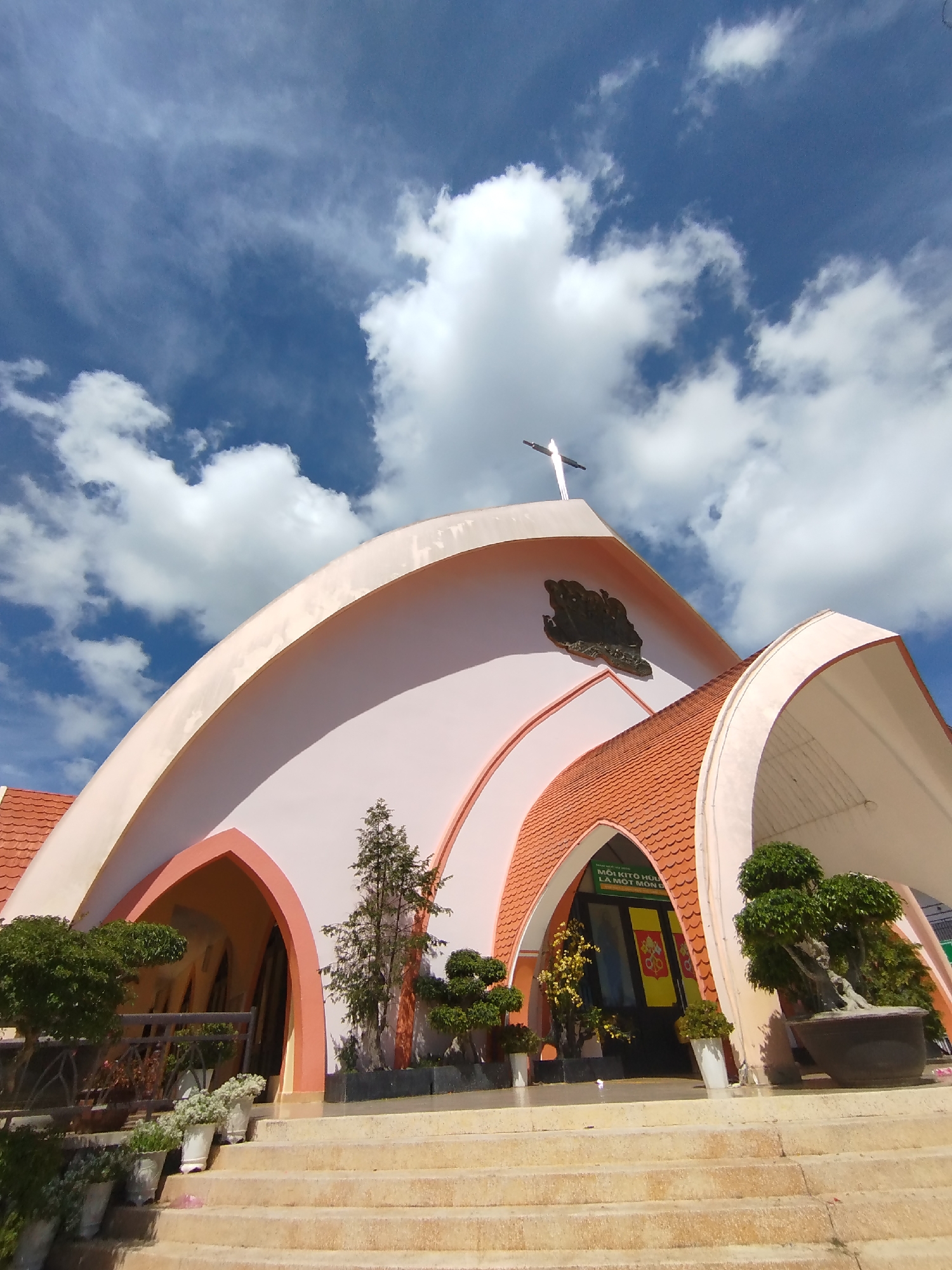 The image shows a low-angle view of a building against a backdrop of a blue sky with scattered clouds. The building appears to be a church or religious structure, characterized by its pink and orange color scheme and arched architectural features. The roof is tiled with reddish-orange tiles, and a white cross is mounted on the highest point of the structure. There are plants and greenery in pots or planters at the base of the building, near a set of stairs leading to the entrance. A sign with text in what appears to be Vietnamese is above the entrance, stating in Vietnamese "Moi Kito Huu La Mot Mon Doc". The location is the Church of Phu Cam, in the city of Da Lat, Vietnam.