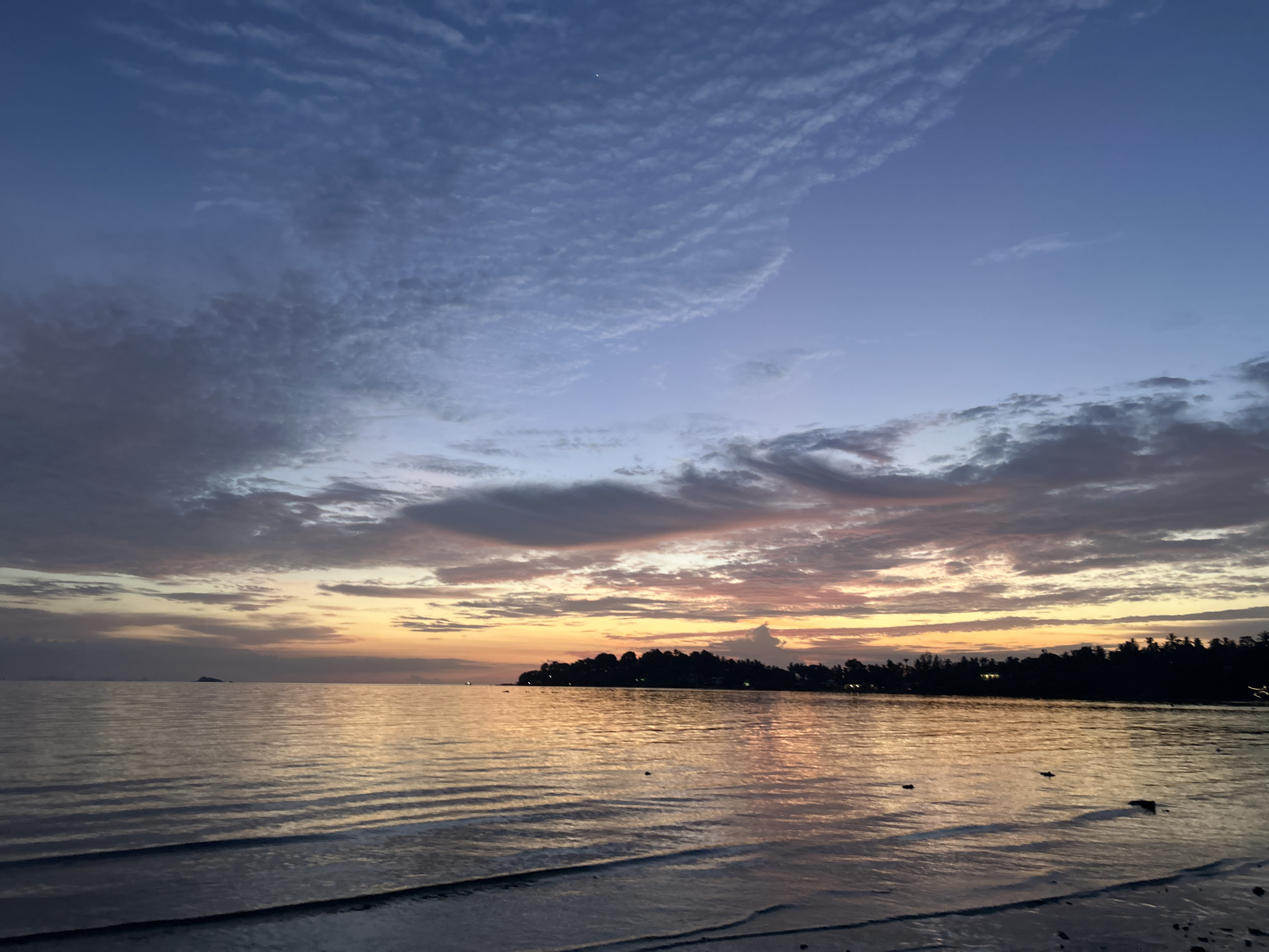 A wide shot captures the tranquil seascape of Ko Pha-ngan District, Thailand, at dusk. The sky transitions from a deep twilight blue at the top to soft hues of pink, orange, and yellow near the horizon, where clouds are painted with the fading sunlight. A thin sliver of moon is visible in the upper left quadrant of the sky. The sea, calm and reflective, mirrors the vibrant colors of the sunset, with gentle waves lapping at the shore in the foreground. A silhouetted treeline, marking the coastline, stretches across the middle ground, punctuated by a few distant lights, suggesting a settlement or village. A small, dark island or structure is discernible on the horizon to the left. The overall atmosphere is serene and peaceful, typical of a beachside sunset in this tropical location.