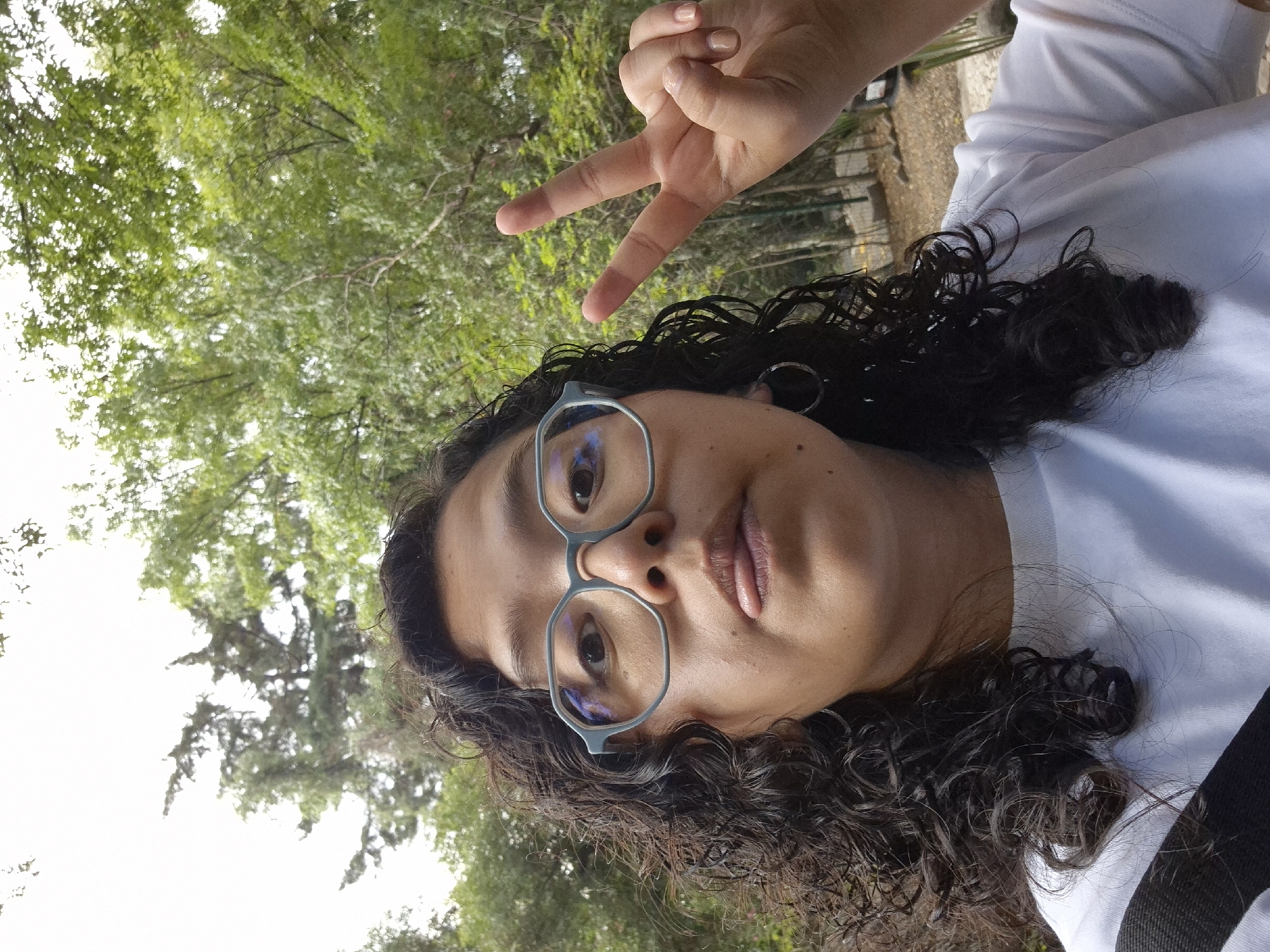 A selfie shot shows a person wearing glasses and making a peace sign, standing in a wooded area. The person has curly dark hair and a light complexion. They are wearing a white shirt with a dark strap visible. The background features green foliage and trees. Based on the foliage type, this image might have been taken in Parque del Centenario in Puebla, Mexico.