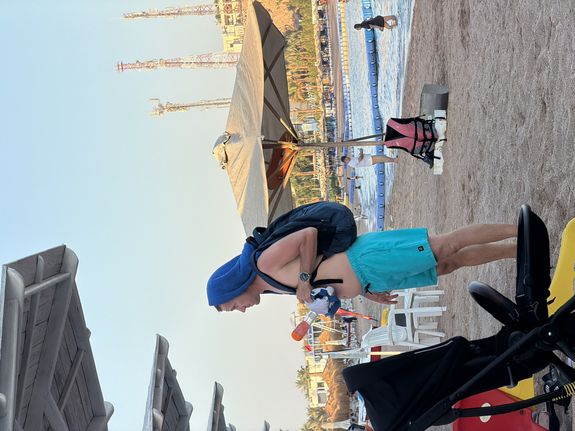 The image depicts a beach scene in Sharm El Sheikh, Egypt, with a man in the foreground wearing blue shorts and a blue towel draped over his head and shoulders. He has a backpack on and is holding a snorkel and mask. The beach is sandy with several beach umbrellas and chairs visible. In the background, the Red Sea is visible, with a roped-off swimming area and several people in the water. There are also tall communication towers in the distance. The lighting suggests it is late afternoon.