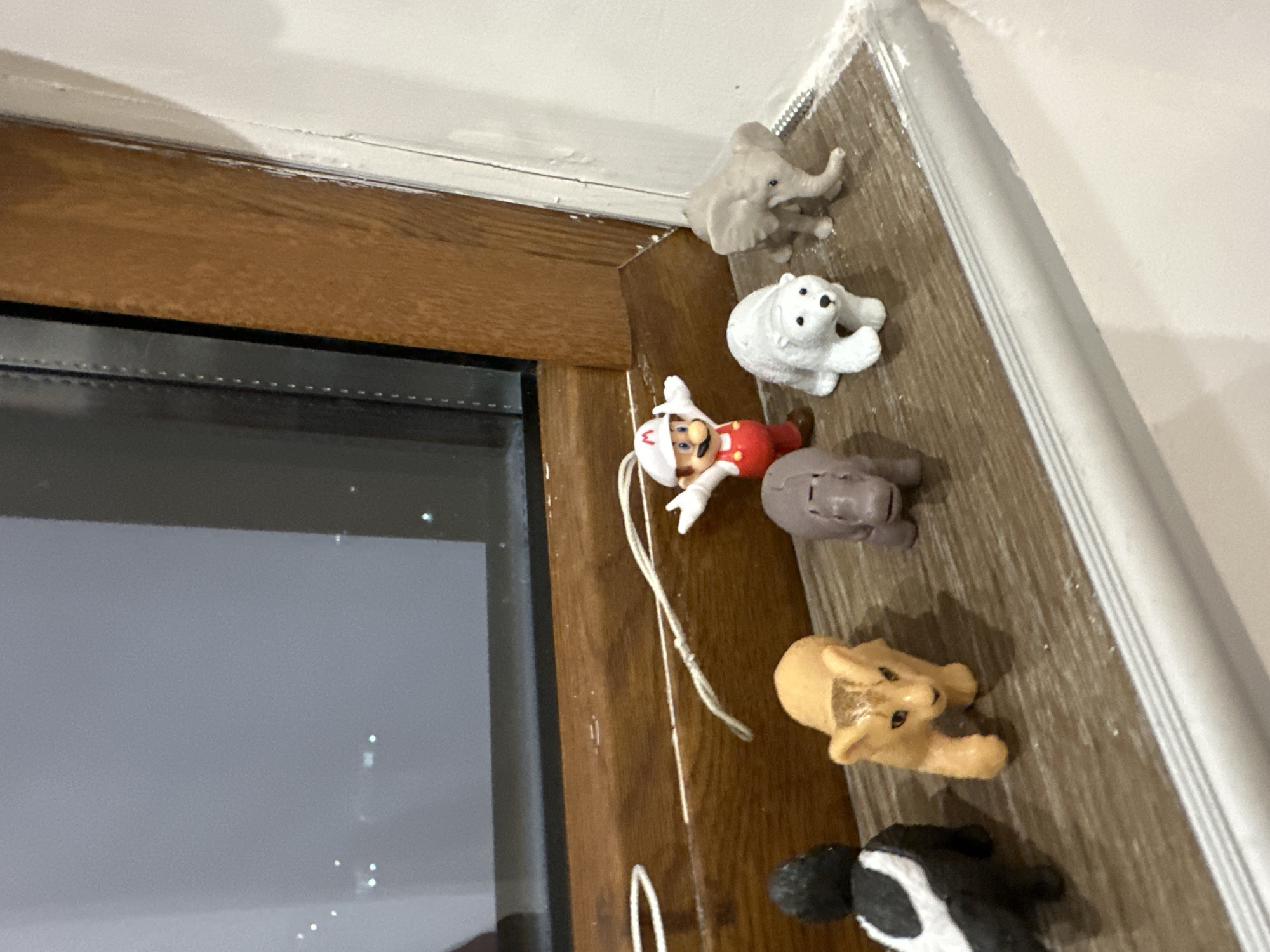 The image shows a collection of miniature toy figures arranged on a wooden surface next to a window. The figures include an elephant, a polar bear, a figure resembling Mario from the Super Mario franchise, a hippopotamus, a lion, and a skunk. The toys appear to be positioned along the edge where the wooden surface meets a white wall. The window is partially visible, reflecting the interior of a room. The image was not taken in Lovech, Bulgaria.