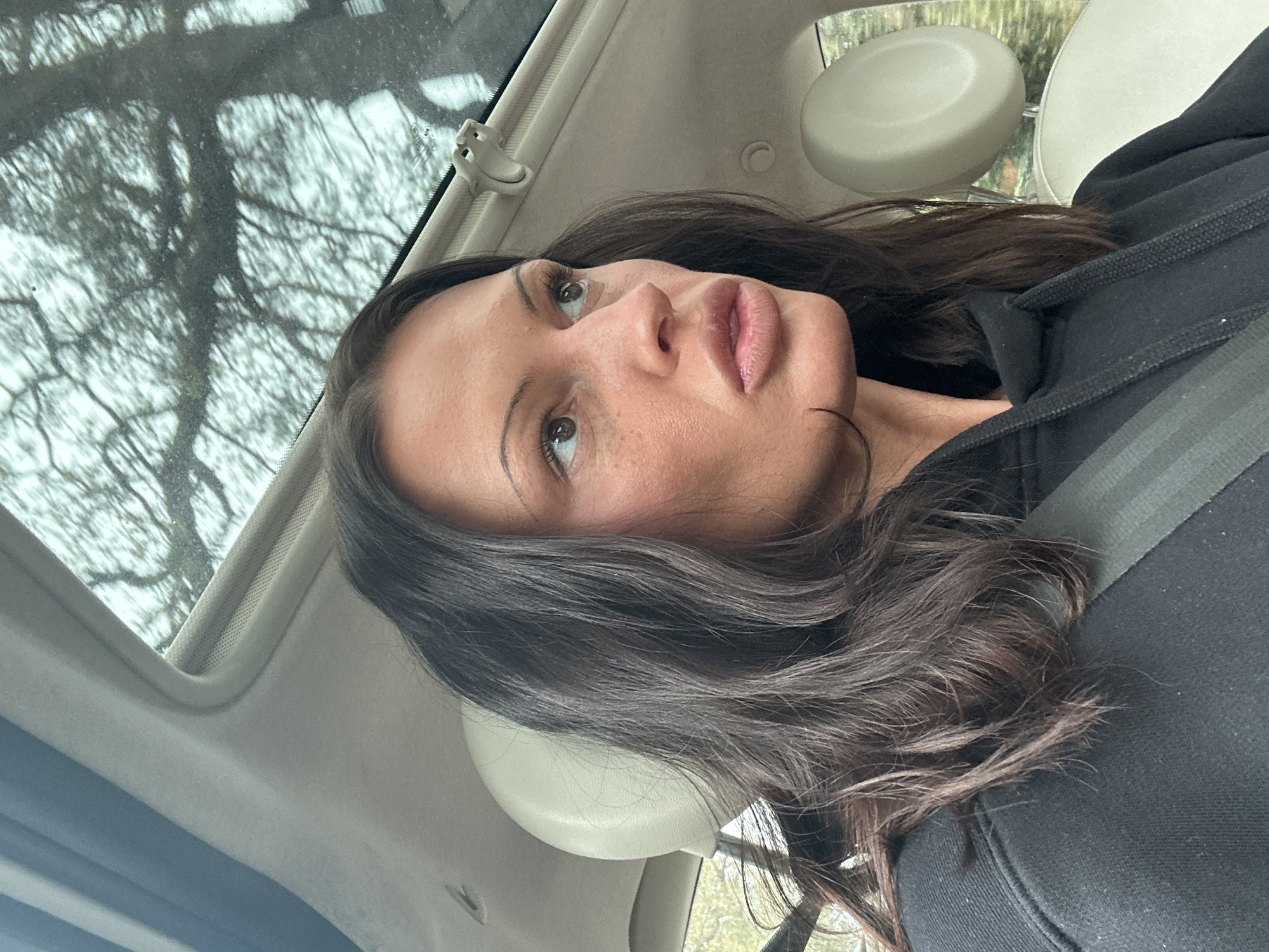 A woman with dark hair is photographed from a low angle inside a car, possibly in Tunbridge Wells, United Kingdom. She is wearing a black top and a seatbelt. The car has a sunroof, which allows a view of trees above. The interior of the car is light in color. The woman's face is in focus, showing her features. The scene is captured under natural lighting.