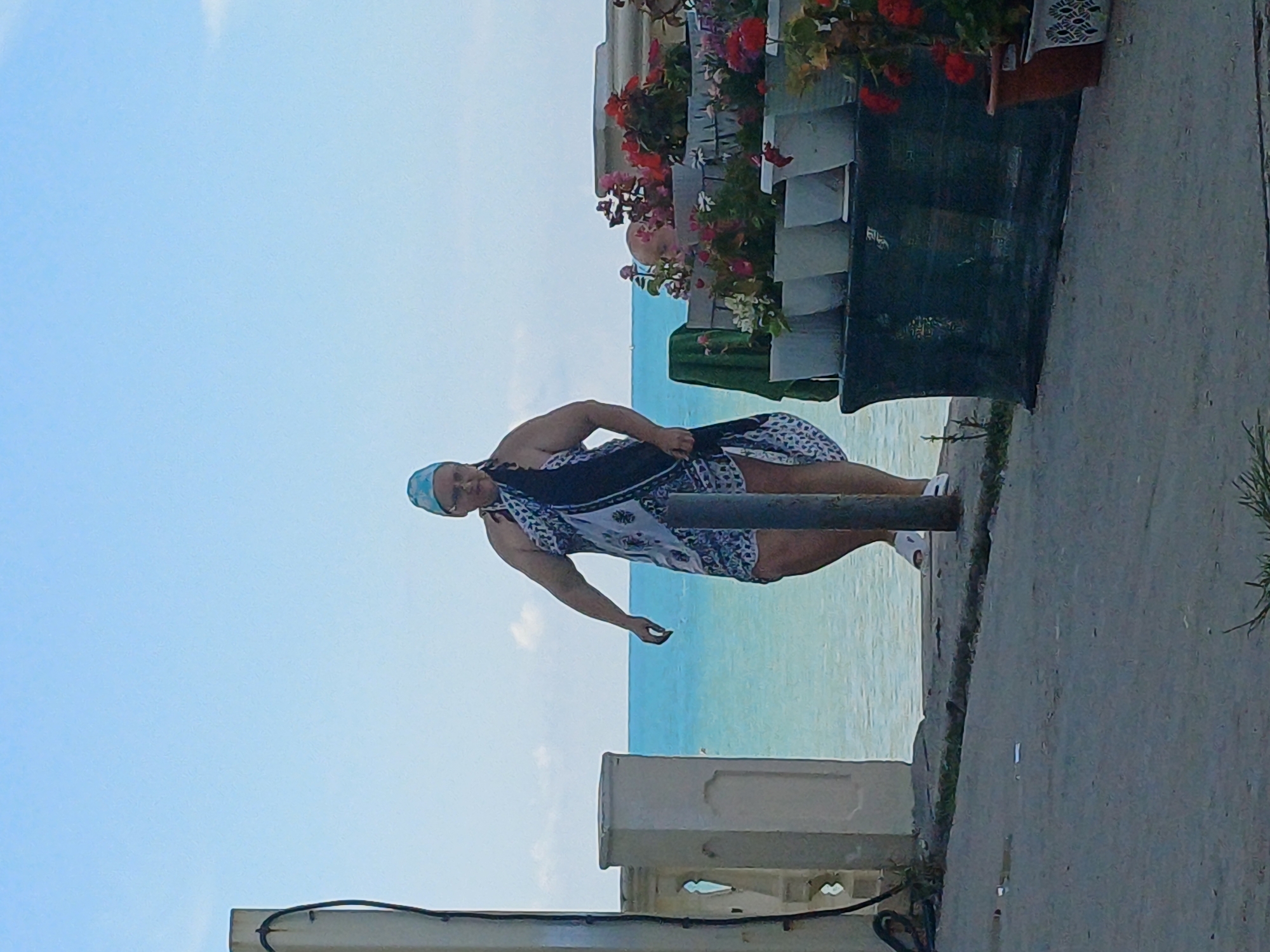 A woman stands near the sea in Sochi, Russia. She is wearing a headscarf and a patterned sarong, and appears to be walking on a paved area. In the background, the turquoise water of the sea meets a clear blue sky. To her right, there are several potted plants arranged on a dark green structure. To her left, a white railing and a pillar are visible. The overall scene suggests a sunny day at the seaside.