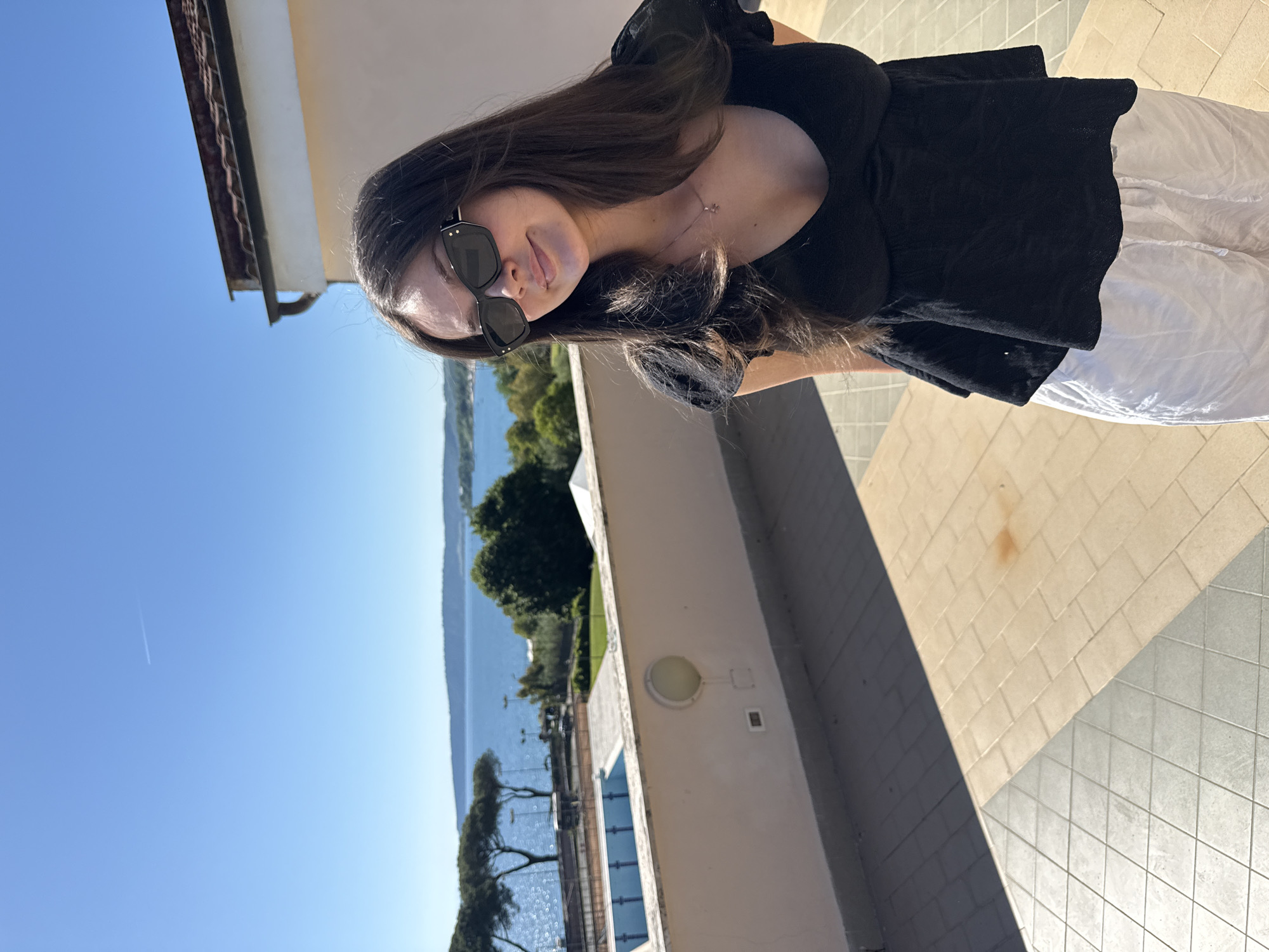 A young woman with long, dark hair and sunglasses stands on a tiled terrace, wearing a black top and white pants. She is oriented towards the camera. The terrace overlooks a body of water, presumably Lake Bracciano, surrounded by green trees and hills. A clear blue sky is visible above. Part of a building with a light fixture is visible behind her. Based on the location, the image was likely taken in or near Bracciano, Italy. A pool is visible beyond the railing.