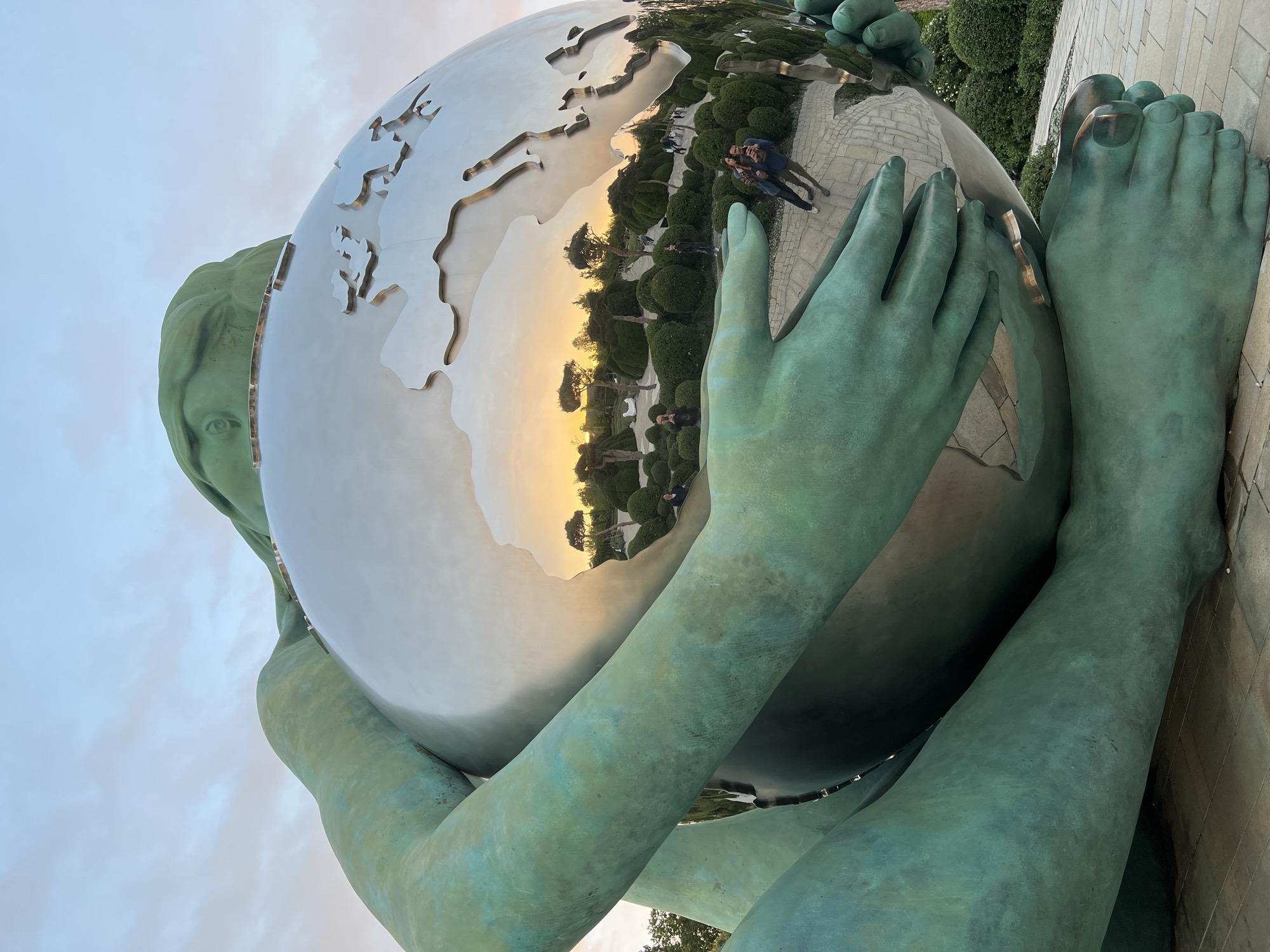 A low-angle, tilted photograph captures a monumental outdoor sculpture, likely located in Krasnodar, Russia. The primary subject is a colossal statue of a human-like figure, appearing to be female, crafted from a material that has developed a distinct teal-green patina, resembling aged bronze or copper. The figure is depicted in a squatting or kneeling posture, cradling a large, highly reflective spherical globe with both hands and arms.
The globe's surface is a polished, mirror-like metal, featuring engraved or raised outlines of continents. This reflective surface prominently displays a clear reflection of the surrounding environment: two individuals are visible, standing on a paved walkway, appearing to pose or observe. Behind them, a carefully landscaped garden with manicured green bushes and trees stretches into the distance. The reflection also reveals a warm, golden glow on the horizon, suggesting late afternoon or early evening light.
Above the statue, the sky is overcast with soft, diffused light. Notable details of the statue include a partially visible eye peeking from behind the globe on its left side, as well as large, stylized hands and feet. Subtle gold-colored accents are present on the statue's feet, possibly indicating jewelry or painted features. The overall scene suggests a well-maintained public park or garden setting. No visible text is present in the image.