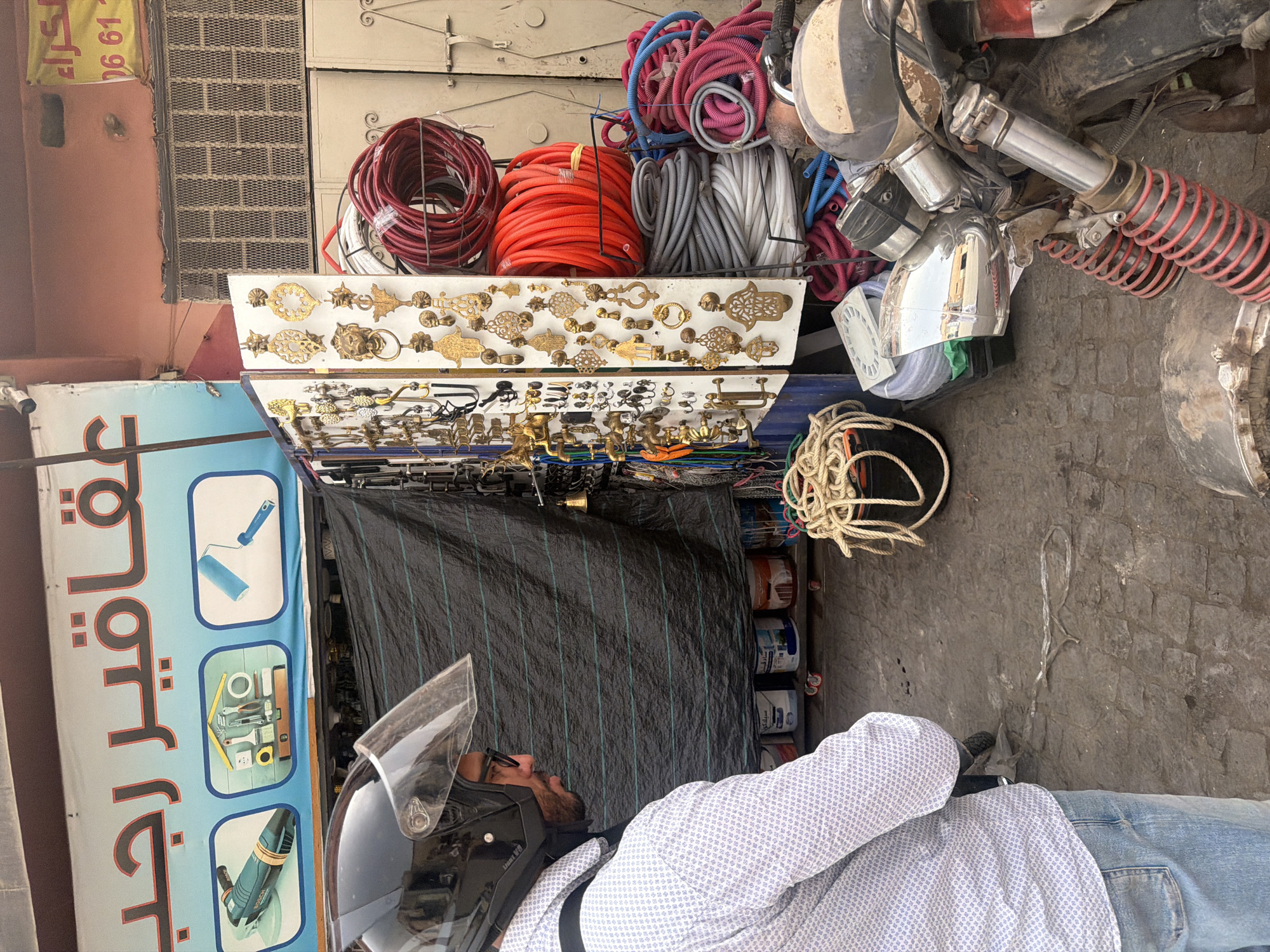 A man wearing a helmet, glasses, a patterned shirt, and jeans is standing in front of a storefront in Marrakesh, Morocco. The storefront displays a variety of items, including metal hardware such as door knockers and decorative pieces, coiled hoses in red, gray, and pink, and buckets of paint. A motorcycle is parked on the right side of the image. The scene appears to be an outdoor market or shop, with a cobblestone ground and a blue banner with Arabic text and images of tools above the storefront.