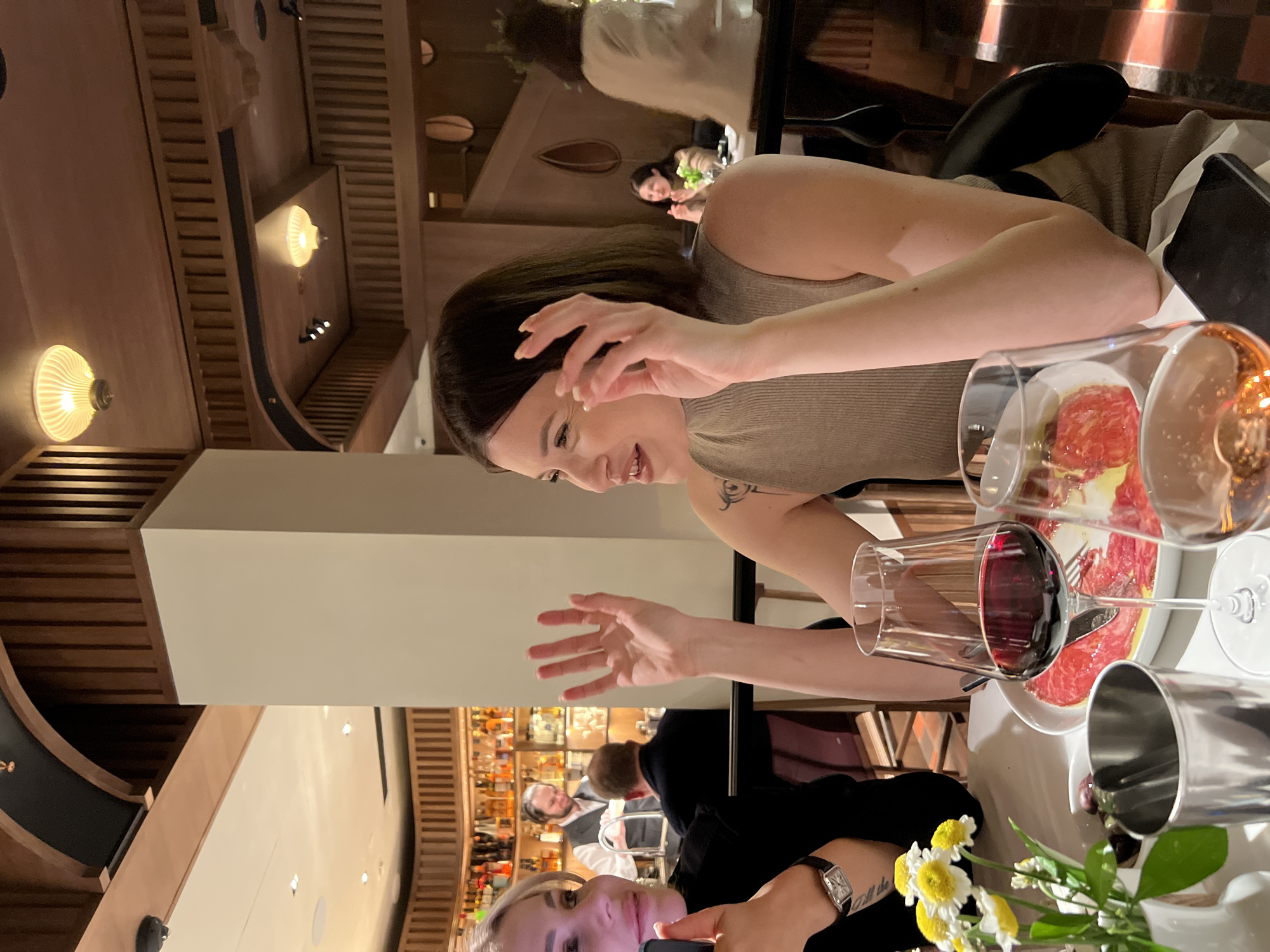 This media file captures an indoor scene in an upscale restaurant or bar during the evening. The primary focus is on two women seated at a table, engaged in a meal and drinks.
In the midground, a woman with dark hair, wearing a beige ribbed top, is smiling broadly and gesturing with both hands, indicating active conversation or excitement. A tattoo is visible on her right arm. In the foreground, partially visible, another woman with blonde hair, dressed in a black top, holds a phone, seemingly capturing a moment. A watch adorns her left wrist, below which a tattoo with the fragmented text "till the" is visible.
On the table between them, a white plate holds a dish composed of thinly sliced red meat, possibly carpaccio, topped with a yellow sauce. Two wine glasses are present, one filled with red wine. A silver container, likely an ice bucket, and a small white vase holding a bouquet of white and yellow daisies complete the table setting.
The background showcases the restaurant's warm and inviting ambiance, featuring extensive wooden slatted panels on the walls and ceiling. Multiple wall sconces with ribbed glass shades emit a warm, yellowish light, setting an evening mood. A well-stocked bar area with bottles and glassware is visible further back, along with blurred figures of other patrons and a bartender. The overall scene conveys a pleasant and social dining experience.