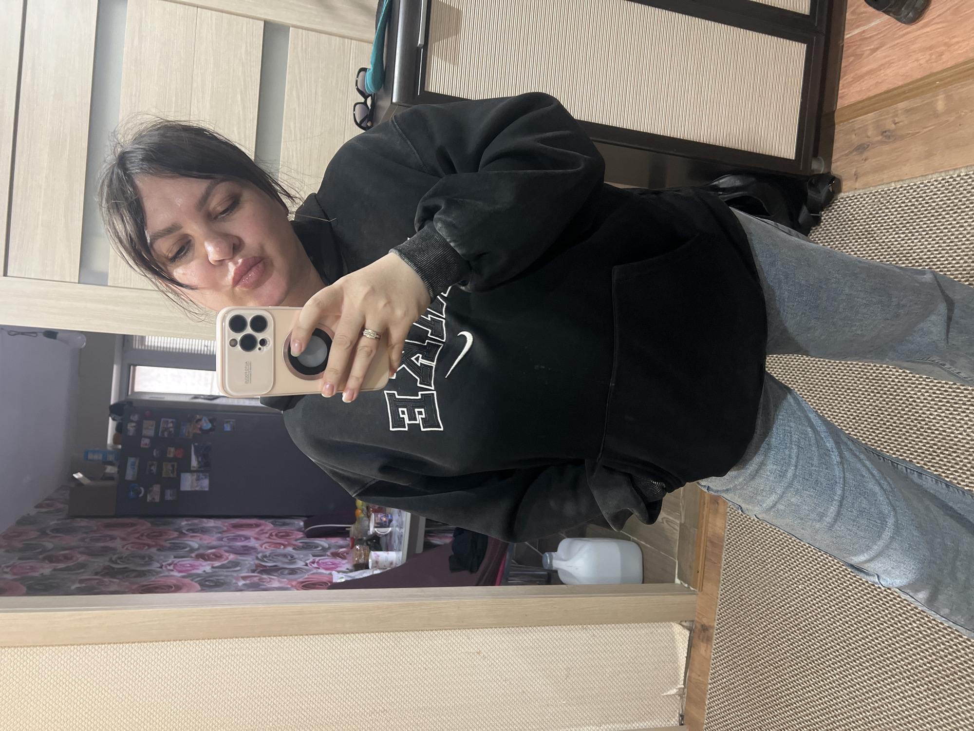 A woman with dark hair is taking a selfie in what appears to be a mirror. She is wearing a black Nike hoodie and light blue jeans. Her phone has a light-colored case. The background includes a closet, a floral-patterned surface, and a floor with a textured rug. A white jug sits on the surface near the mirror. There is no way to determine if the location is in Noviy, Russia.