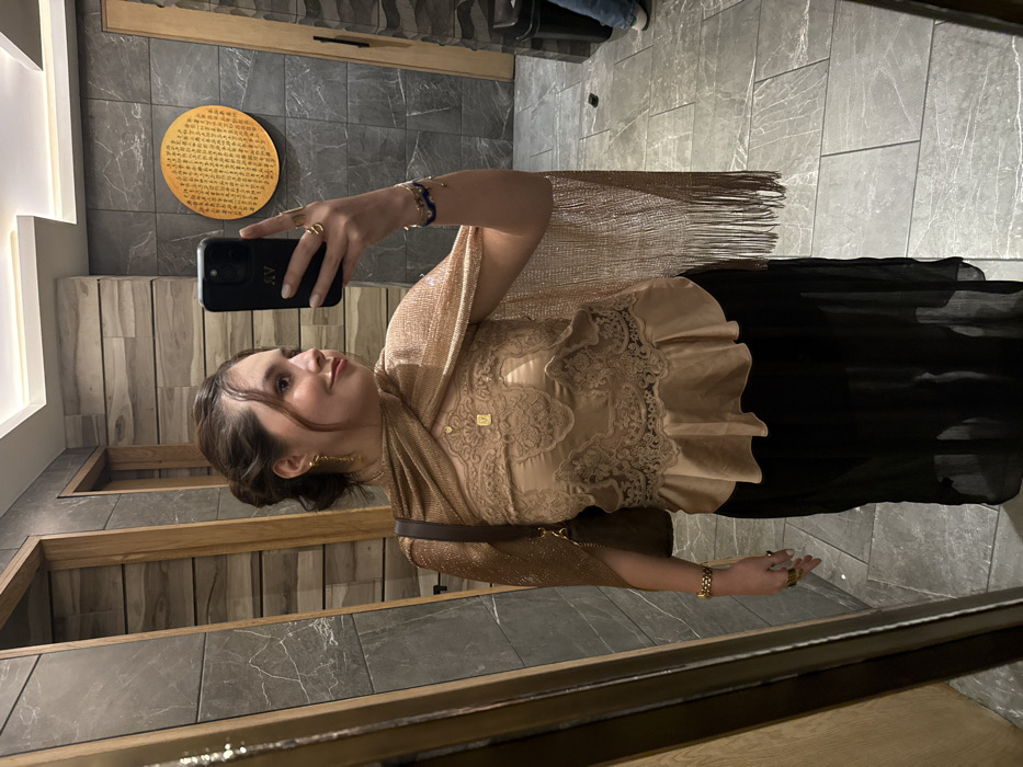 A woman takes a selfie in a mirror. She wears a gold, lace-trimmed top and a black skirt or dress. She has a gold shawl with fringe over her shoulder and several bracelets on her arm. A small gold pendant hangs from her neck. The background shows a bathroom with gray tiled walls and a decorative wall hanging. A wooden frame surrounds the mirror.
