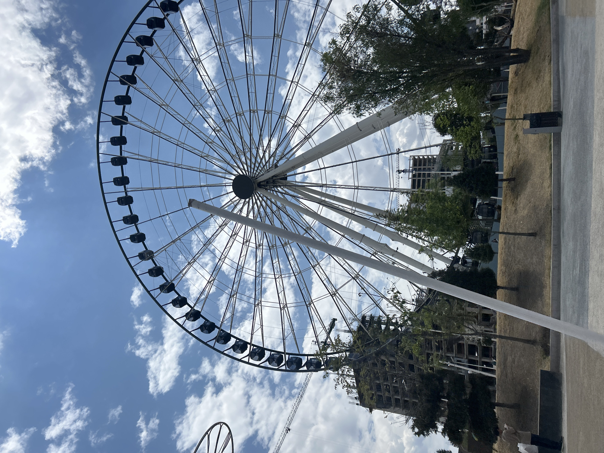 The image shows a Ferris wheel against a sky with scattered clouds. The wheel is primarily black with a network of white supports and gondolas. In the background, there is a partially constructed building and some trees. To the right, there is a grassy area and a concrete walkway. The image suggests an urban park or recreational area. It's a daytime scene with good visibility. Given the prompt, it is presumably located in San Bernardino Tlaxcalancingo, Mexico.
