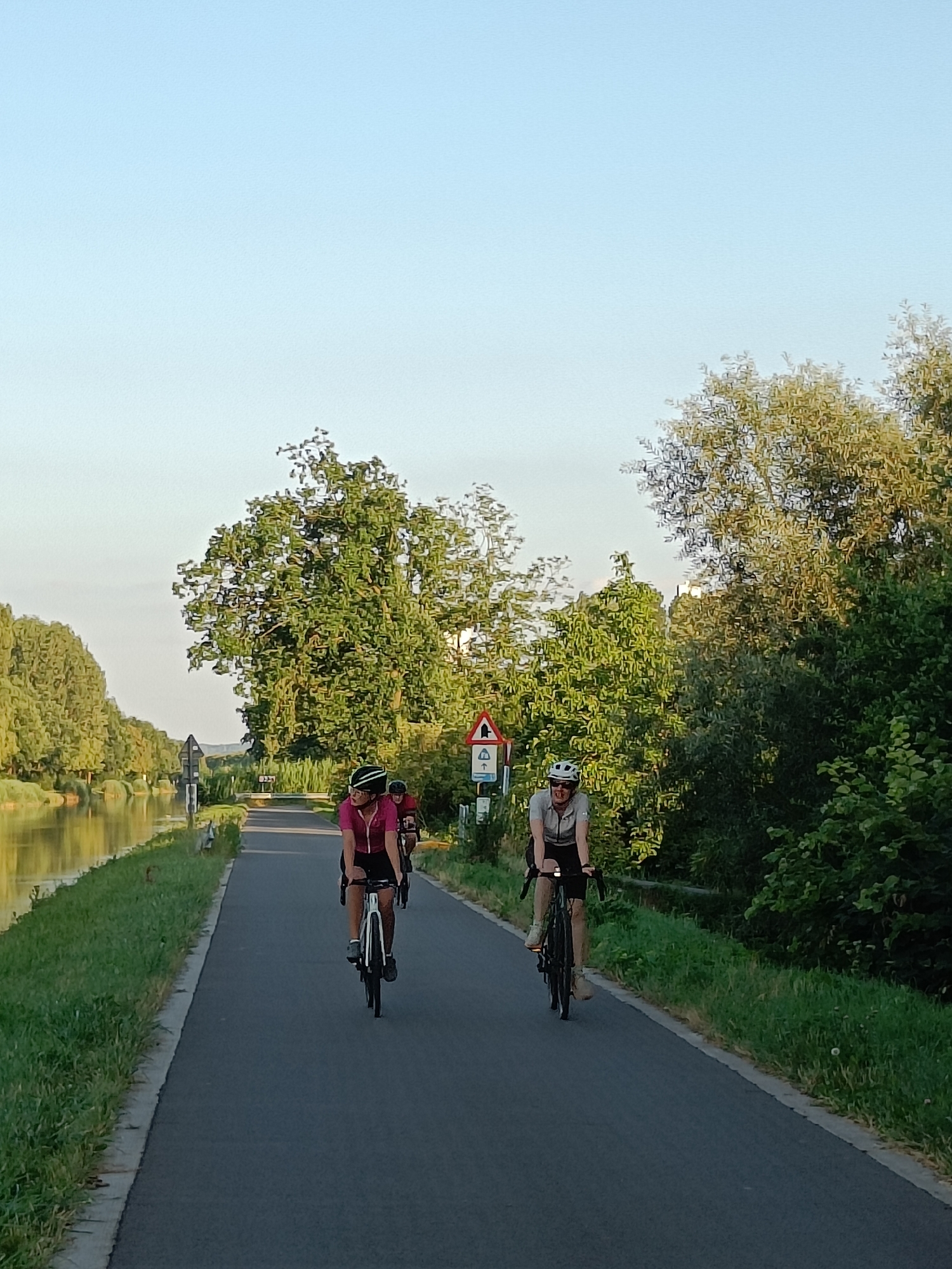 Two individuals on bicycles ride along a paved path next to a canal. The person in the foreground is wearing a pink shirt and a helmet, with a black bicycle. Behind them, another person on a bicycle is visible, also wearing a helmet. The path is bordered by grass and trees on both sides, with the canal on the left. A traffic sign is visible on the right side of the path. The sky is clear and blue. The location is the canal in Haacht, Belgium.