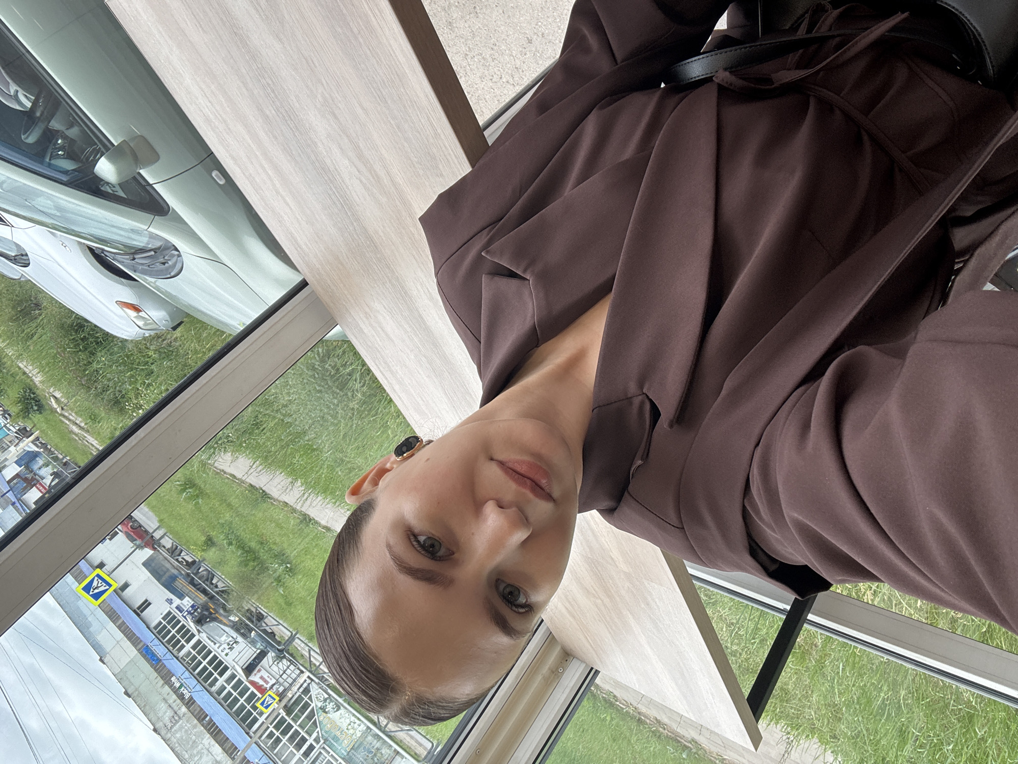 A young woman with her hair pulled back and wearing a brown blazer takes a selfie indoors, seated at a wooden table by a window. Through the window, a street scene is visible in Chaikovsky, Russia, featuring buildings, parked cars, a truck, and greenery. The woman is wearing black earbuds.