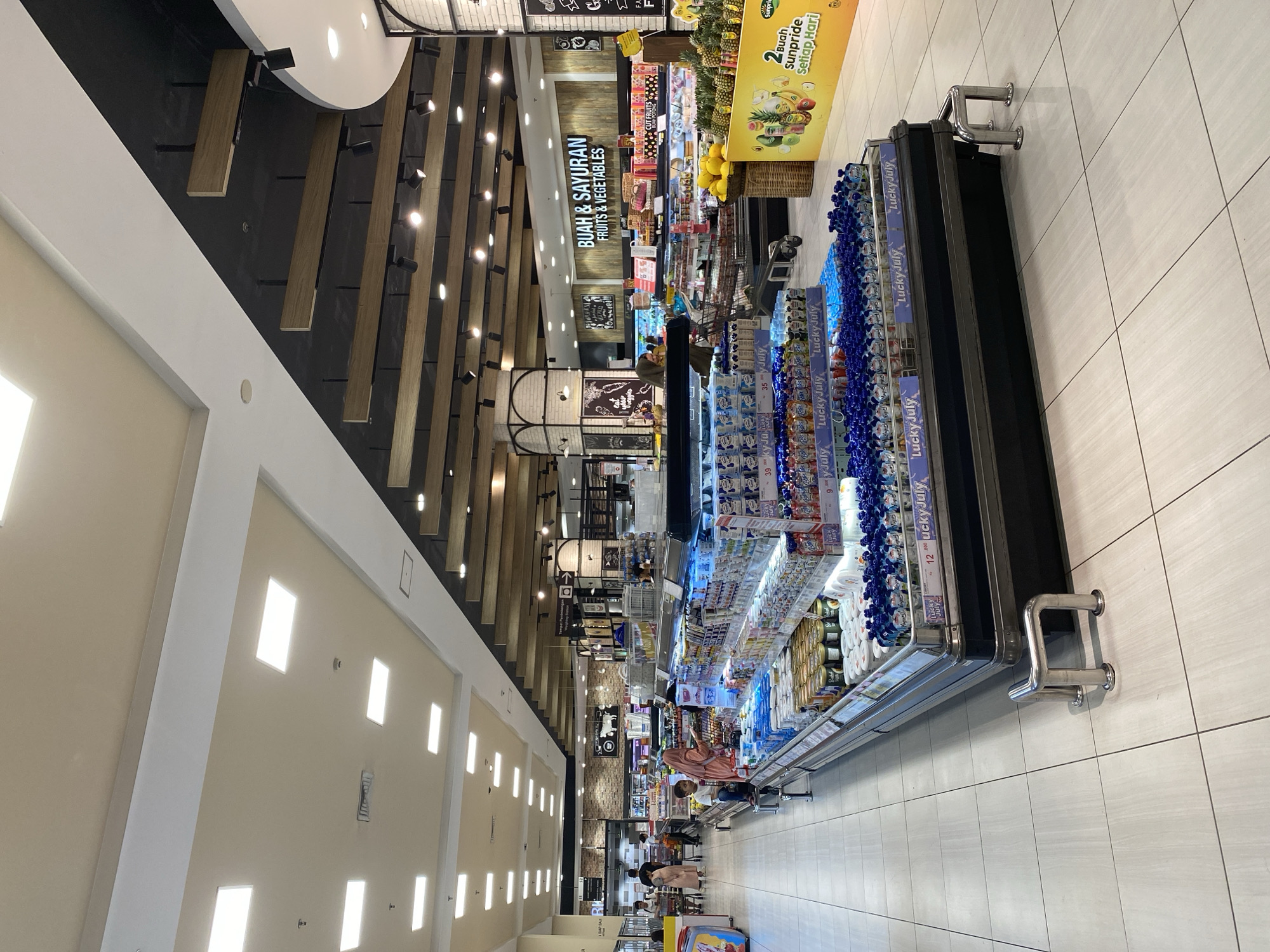 The image depicts the interior of a brightly lit supermarket in East Jakarta, Indonesia, likely a brand like "Super Indo" based on the signage. The scene focuses on a refrigerated aisle filled with a wide assortment of dairy products, including yogurt and milk drinks, predominantly branded "Lucky July." These items are neatly arranged on multiple shelves within a long, glass-fronted display case. In the background, shoppers are visible browsing the aisles, with some pushing shopping carts. Further back, signs indicate sections like "BUAH & SAYURAN" (Fruits & Vegetables). The flooring is tiled in a light beige color, and the ceiling features a modern design with recessed lighting and wooden accents. The overall atmosphere is clean and well-organized, typical of a modern grocery store.