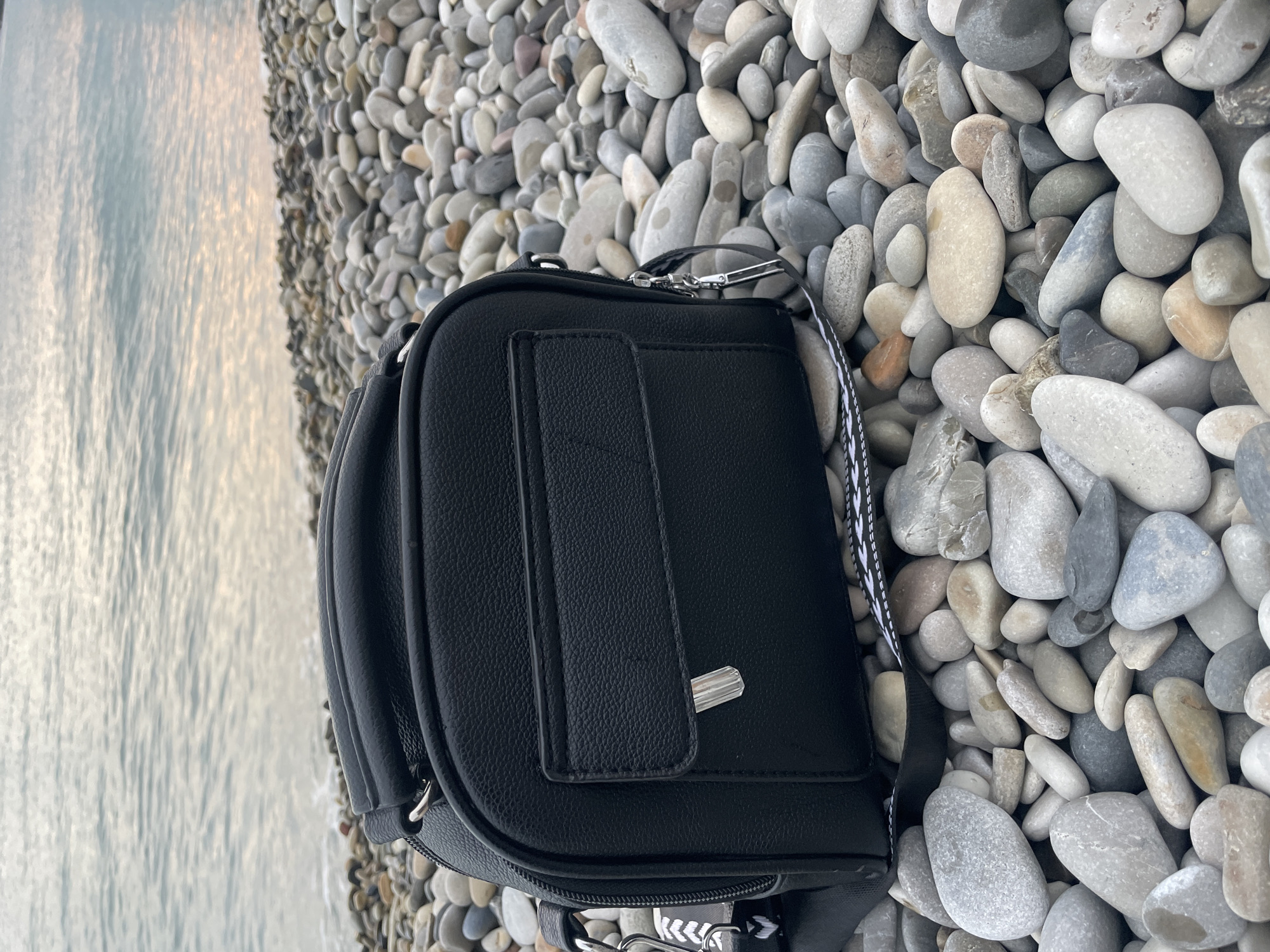 The image shows a black leather bag lying on a pebble beach in Olginka, Russia. The sea is visible in the background. The pebbles are mostly gray and white. The bag has a rectangular shape with a flap closure. A strap with a black and white pattern is attached to the bag.